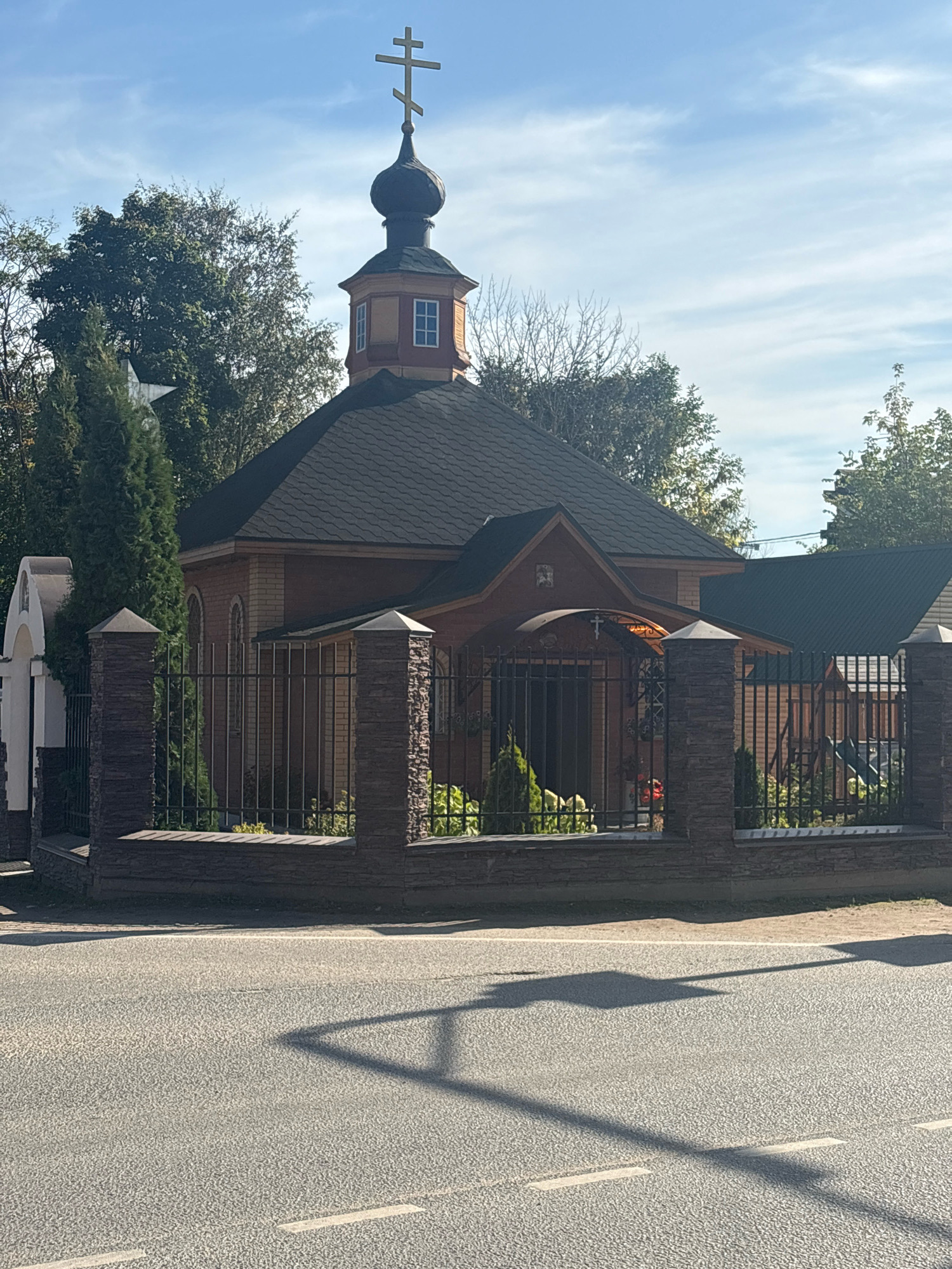 The image shows a small, wooden Orthodox church with a dark roof and a bulbous dome topped with a cross. The church is surrounded by a black metal fence with stone pillars. There are trees and greenery behind the fence and to the left of the church. A white structure with a cross is visible to the far left. The foreground shows an asphalt road with white markings and a shadow cast by a street light or pole. The sky is blue with scattered clouds. The image is taken outdoors during the daytime.