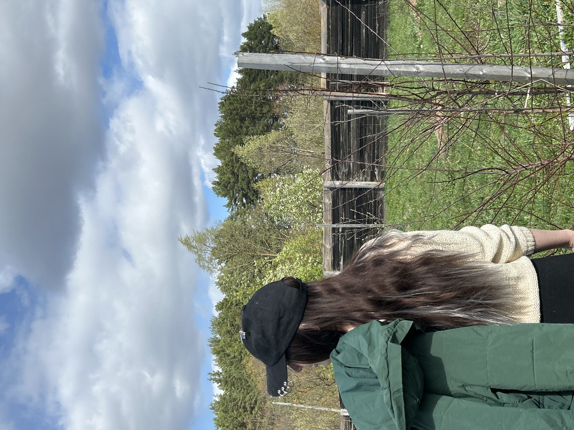 The image depicts a person wearing a black baseball cap, a light-colored sweater, and black leggings, standing outdoors on a grassy area. A green jacket is draped in front of them. Behind them is a wooden fence and trees with some bare branches. The sky above is partly cloudy with patches of blue visible. The location is in a rural area, and there is no discernible evidence to confirm the specific location of Rusalevka, Russia.