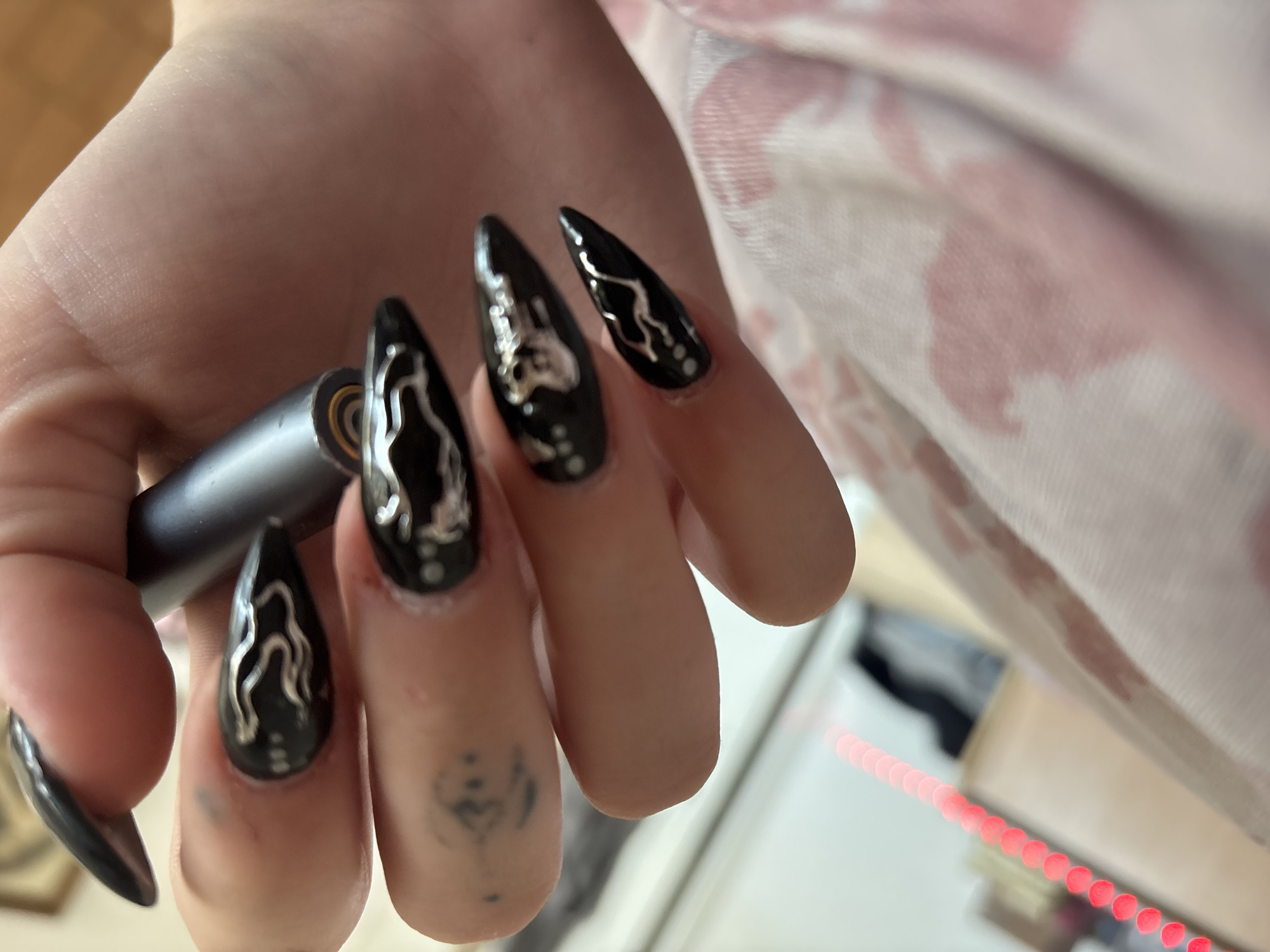 A close-up shows a hand with long, black, pointed nails adorned with silver designs, holding a cylindrical object. The background is blurred, showing a pink and white patterned surface, a light-colored wall, and a strip of red lights. A tattoo is visible on the thumb. The location is not discernible from the image.