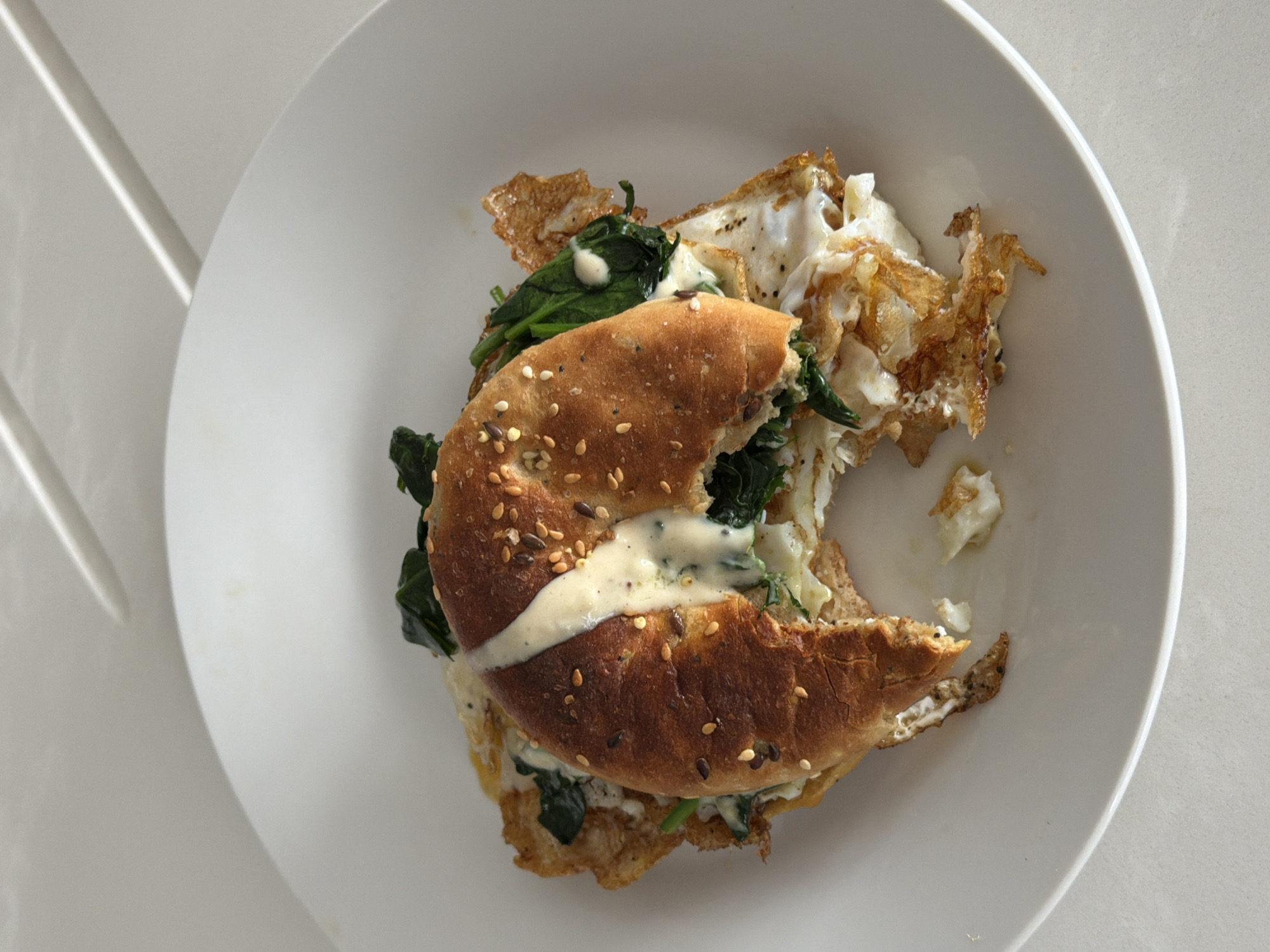 The image shows a half-eaten bagel sandwich on a white plate. The bagel appears to be multi-grain, with sesame seeds. The filling consists of cooked spinach, a fried egg, and a creamy white sauce. The egg's edges are crispy. The plate sits on a white surface. A reflection of light appears in the upper left corner. There is no evidence to indicate this scene's location in the city of London, United Kingdom.
