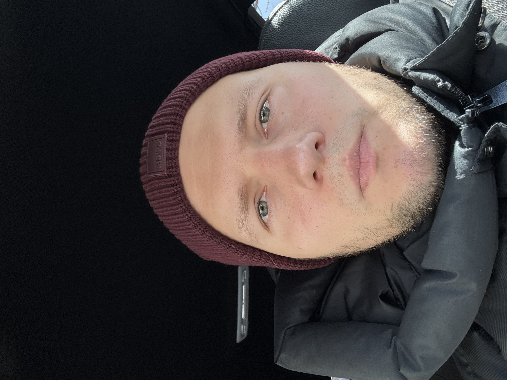 A close-up, slightly angled selfie depicts a fair-skinned male with short blonde hair, a light beard, and green eyes. He is wearing a maroon beanie with a logo patch on the left side. He is also wearing a dark-colored puffy jacket. The background is predominantly black and appears to be the interior of a vehicle, possibly a car, with a portion of the seat visible in the upper center. There is no evidence to suggest the image was taken in Krasnoyarsk, Russia.