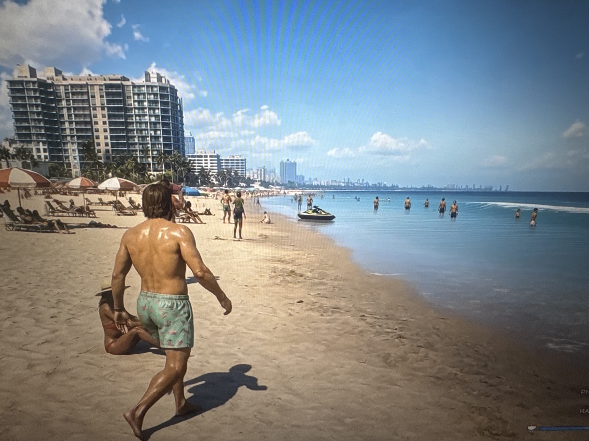 The image depicts a sunny day on a sandy beach with a calm blue ocean. In the foreground, a man with tanned skin and dark hair is walking on the sand, wearing patterned swim trunks. To his left, a woman wearing a hat and a bikini is sitting on the sand. Further down the beach, several people are visible, some lounging on chairs under umbrellas, while others are in the water. In the background, a large apartment building with numerous balconies dominates the coastline, with other buildings and palm trees suggesting a city setting. A jet ski is present in the shallow water. The sky is blue with scattered white clouds. There is no visual information in the image that suggests this scene is located in Stepnogorsk city administration, Kazakhstan.