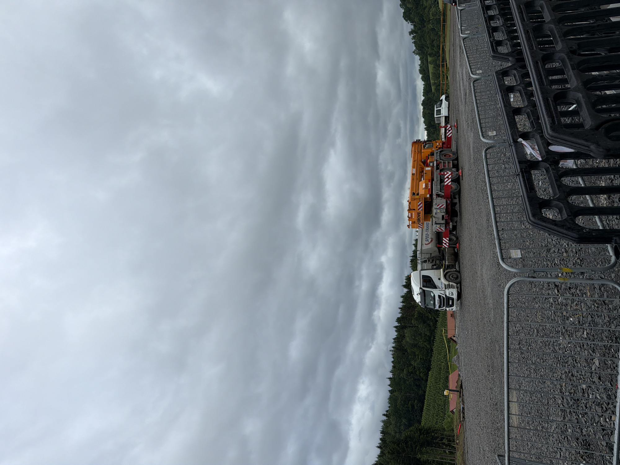 An outdoor, overcast, daytime scene shows a construction or industrial area in Janowice, Poland. In the mid-ground, a white Iveco truck is parked on a gravel surface. To the right of the truck is a large, orange crane. Further back, a white van is visible. A wooded area with a dark green forest lines the horizon behind the vehicles. In the foreground, black and silver barricades are set up. The ground is covered in grey gravel. The sky is heavily clouded, with a pale grey hue.