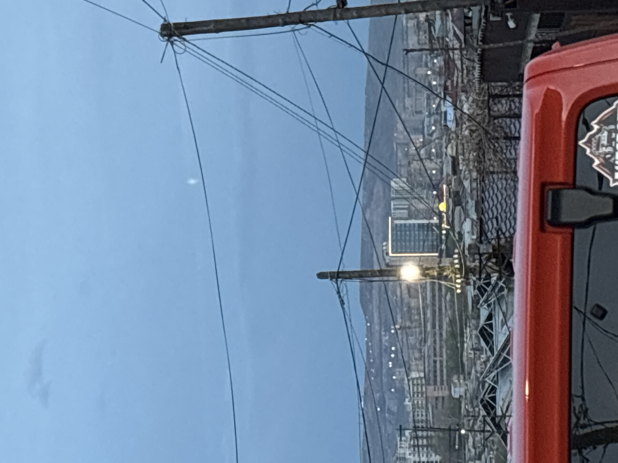 The image captures a cityscape of Yerevan, Armenia, framed by overhead power lines and a portion of a red vehicle on the right. The sky is overcast, casting a muted light over the scene. The city extends toward the horizon, marked by a series of hills. A prominent high-rise building is visible amidst the urban sprawl. Streetlights are interspersed throughout the cityscape, adding to the sense of depth. The red vehicle occupies a significant portion of the frame, obscuring some of the urban details.