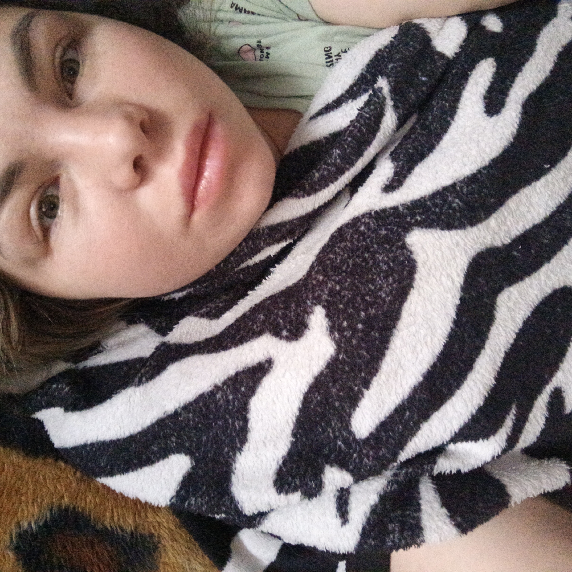 A person lies down with their head angled toward the upper-left of the frame. The person's face is in focus, showing light skin, brown eyes, and glossy lips. A patterned blanket with a zebra print drapes over the body. Parts of a patterned surface, appearing to be animal print, are visible in the bottom-left corner. The background shows a glimpse of a light green garment. No city or location is depicted.