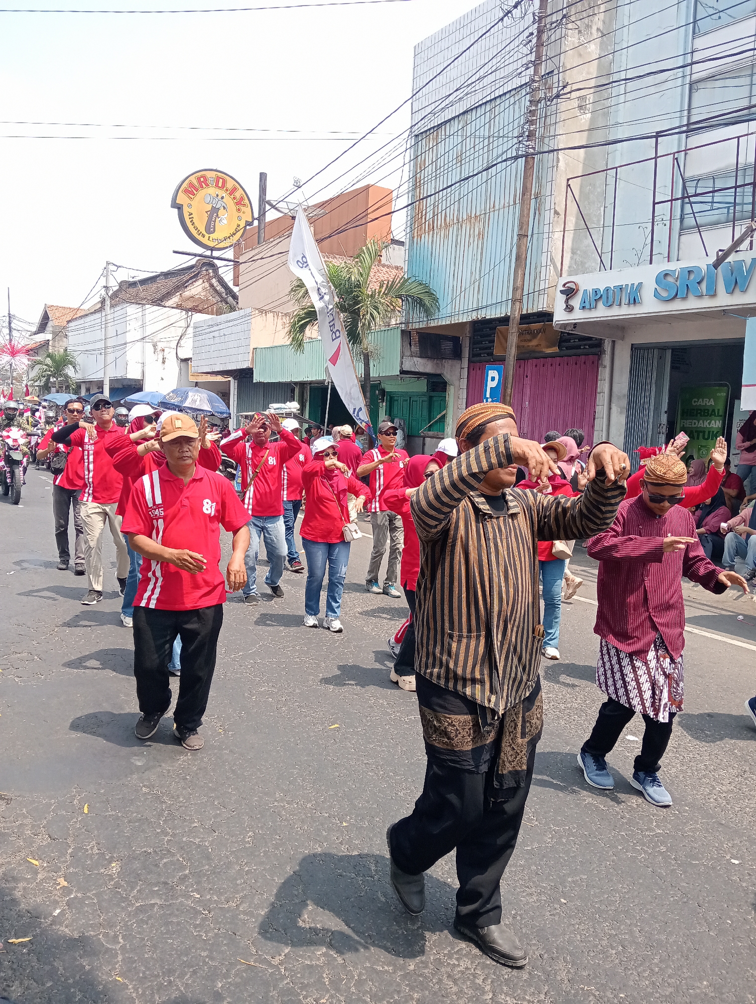 The image depicts a street scene in Demak, Indonesia, where a group of people, predominantly men, are participating in a procession or parade. Many of the individuals are wearing bright red shirts with white accents and the number "81" on them. Some are also wearing traditional Javanese attire, including patterned sarongs and peci hats. The individuals are in motion, with some appearing to be dancing or performing coordinated movements. In the background, there are buildings lining the street, including a store with signage that reads "APOTIK SRIWI." A white flag with Indonesian text is also visible, along with several parked motorcycles and a car. The overall atmosphere suggests a community event or celebration taking place outdoors on a sunny day.