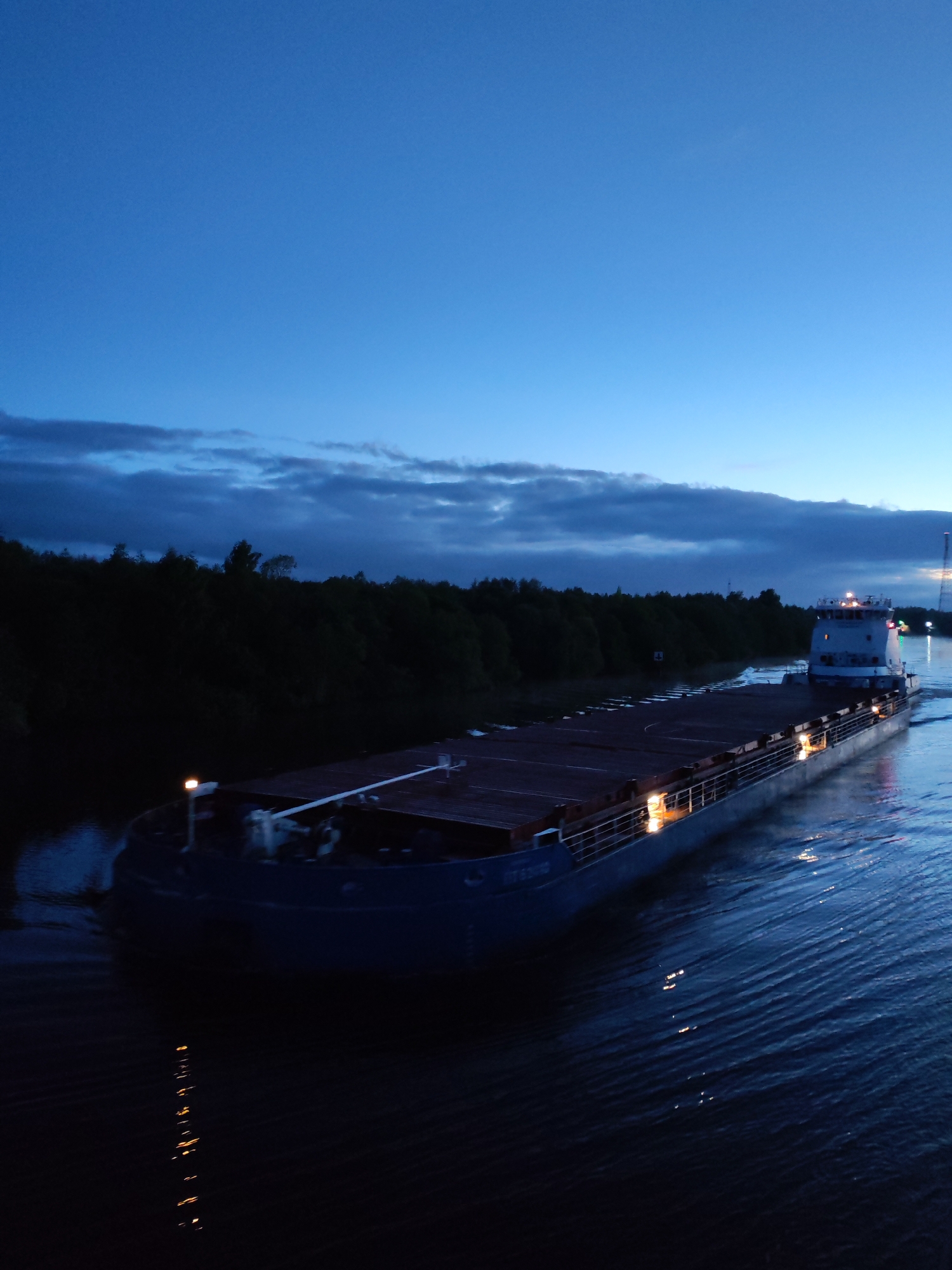 A long, dark barge is moving through a waterway at dusk, likely a canal or wide river. The barge, loaded with a dark cargo, is positioned diagonally in the frame. Small lights illuminate its sides. Dense, dark trees line the banks of the waterway in the background. The sky is a gradient of deep blues, with darker clouds on the horizon. The water surface reflects the lights of the barge and the sky. The location is potentially Annensky Most, Russia, based on the waterway.