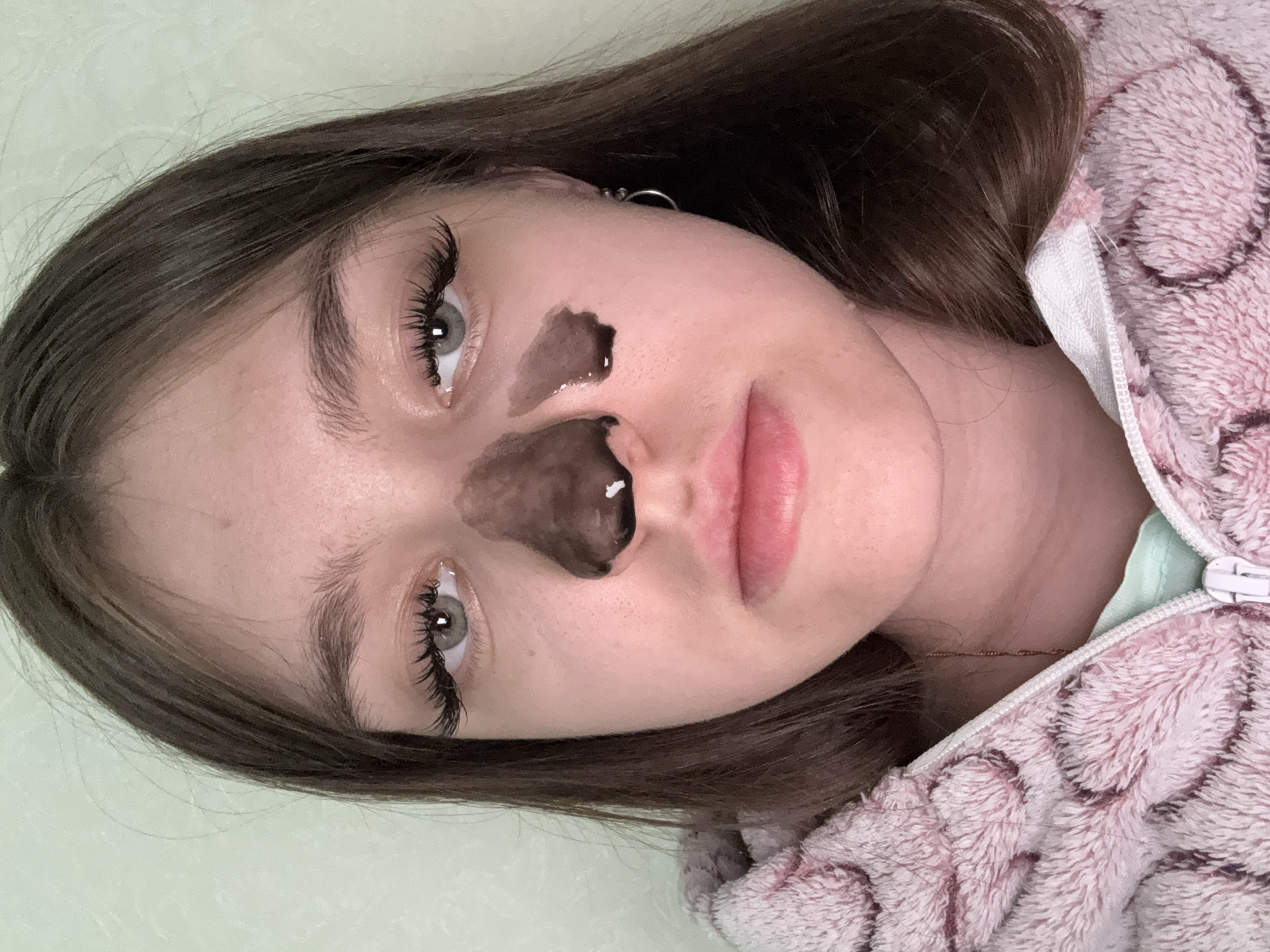 A close-up, vertically oriented portrait features a young woman with long, dark brown hair, fair skin, and light-colored eyes (appearing grey or blue). She is looking directly towards the viewer with a neutral, calm expression.
On her face, she has applied a black peel-off mask, specifically covering her nose and the area between her eyebrows on her forehead. Her long eyelashes appear to be enhanced, possibly with mascara or extensions. Her lips are naturally pink and slightly glossy. A small, light-colored hoop or stud earring is visible in her left ear.
She is wearing a pink, fluffy hooded garment, likely a robe or loungewear, which has a white zipper visible on the right side of the frame. The background is a plain, light-colored surface, possibly a wall, suggesting an indoor setting. The lighting is soft and even. There is no visible text in the image. The overall impression is one of a casual skincare routine.
