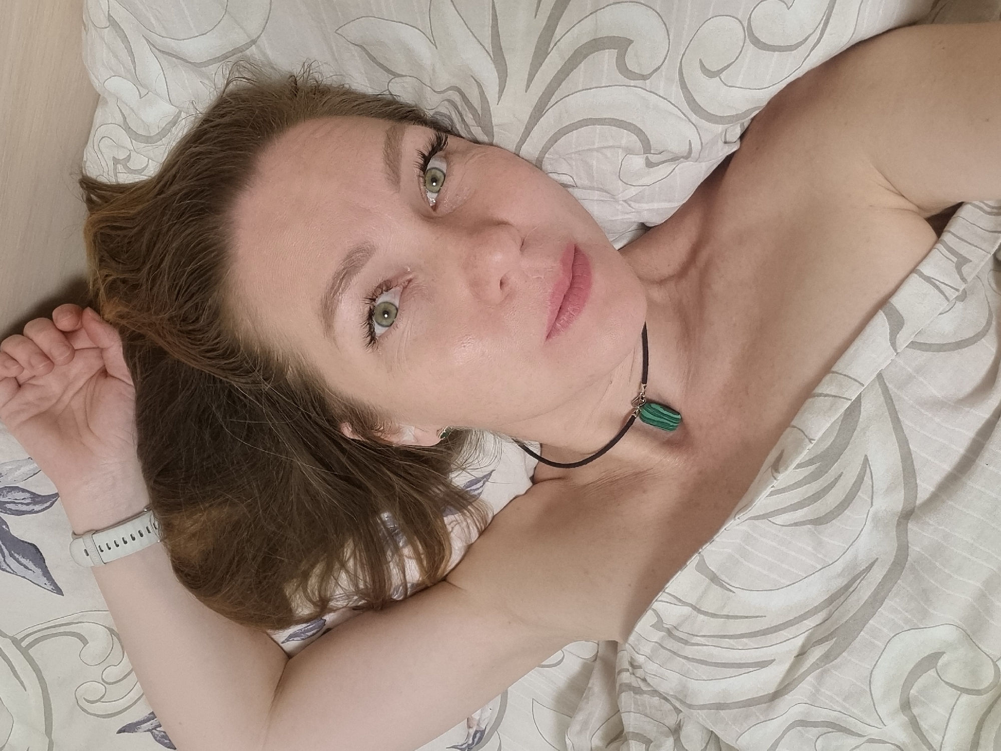 A woman with light skin, light brown hair, and green eyes lies on a bed with white and gray patterned bedding. She wears a black necklace with a green pendant and a white smartwatch on her left wrist. The woman's head is turned towards the camera, and she gazes directly at the viewer. The environment appears to be a bedroom, with a light-colored wooden surface visible near the edge of the bed. There is no indication of the image being taken in the city of Chaikovsky, Russia.