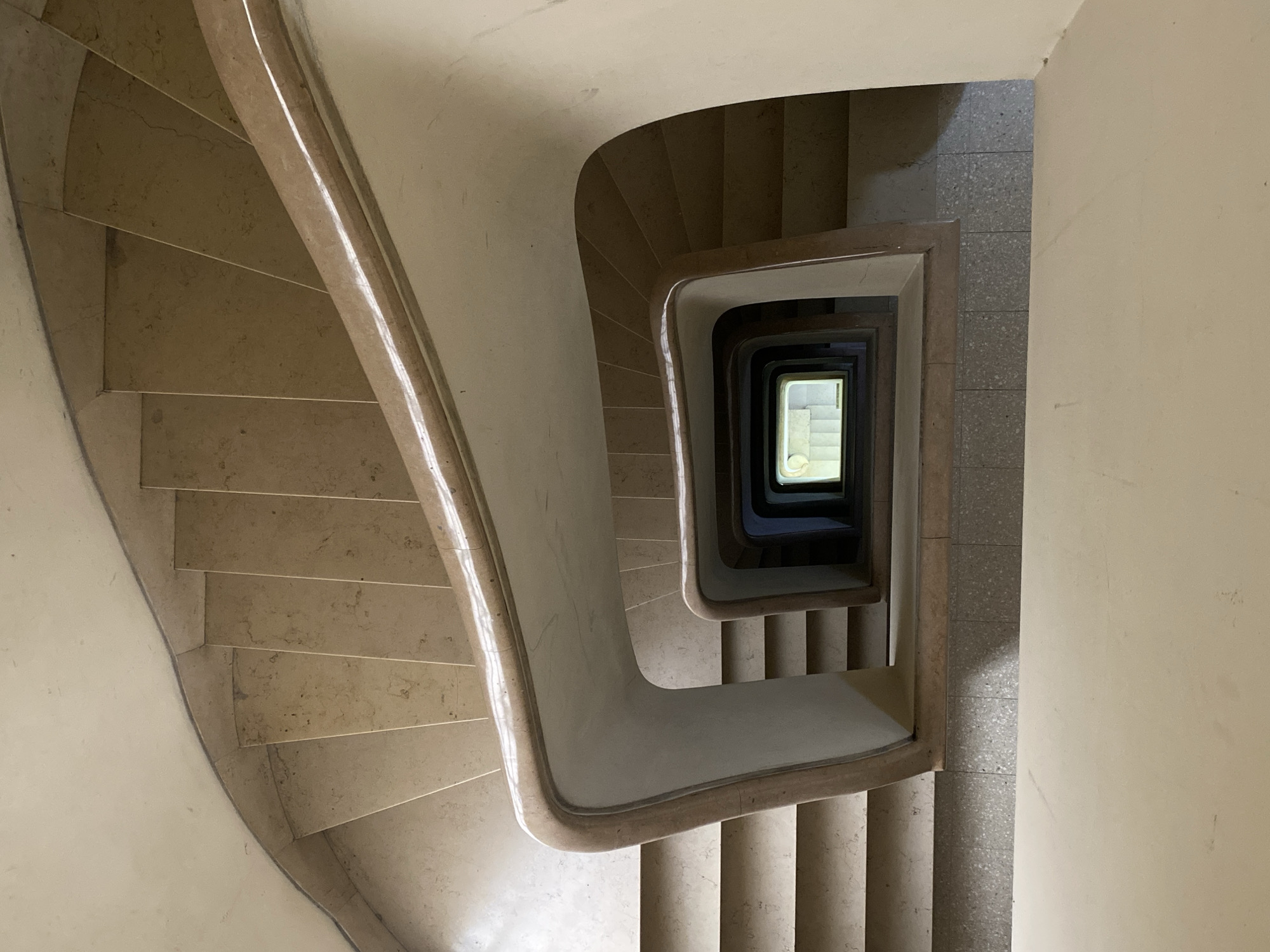 The image provides an aerial perspective looking down a multi-story, square-shaped spiral staircase within a building interior. The staircase and its continuous balustrade are crafted from a light beige or off-white stone or marble, displaying subtle variations in tone and texture. The handrail follows the smooth, curved contours of the balustrade, which features a solid design rather than individual spindles. The walls adjacent to the staircase are a uniform, light cream color.
In the center of the stairwell, a deep, rectangular shaft descends, showcasing the repetitive pattern of the floors below. At the very bottom of this central void, a small, brightly illuminated rectangular opening is visible, appearing to be a window or a light source, revealing a glimpse of another internal space or the outside. This creates a strong sense of depth and a hypnotic, winding effect.
Lighting appears to be natural, possibly from overhead or external windows, casting soft shadows beneath the stair treads and the underside of the balustrades, highlighting the architectural forms. A section of floor to the right of the stairwell is visible, composed of small, light grey, speckled tiles, possibly terrazzo. The scene is devoid of people or any visible text. It's a static architectural shot emphasizing form, light, and depth, in what is contextually located in Buenos Aires, Argentina.