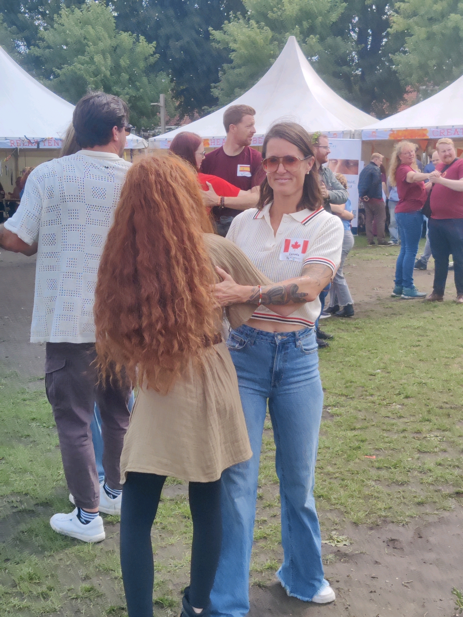 The image captures an outdoor event in Tilburg, Netherlands, with white tents in the background. In the foreground, a woman with auburn, curly hair is facing away from the camera, her arm around the waist of a woman in the foreground. The latter woman, wearing a white polo shirt with red and blue stripes on the collar and sleeves, blue jeans, and sunglasses, is facing the camera and smiling. She has a tattoo on her left arm and a Canadian flag badge on her shirt. To the left, a man in a white, patterned shirt and dark pants is visible from the back, facing the auburn-haired woman. In the middle ground, several other people are mingling, some wearing red tops, and one woman is wearing blue jeans. The ground is a mixture of grass and dirt.