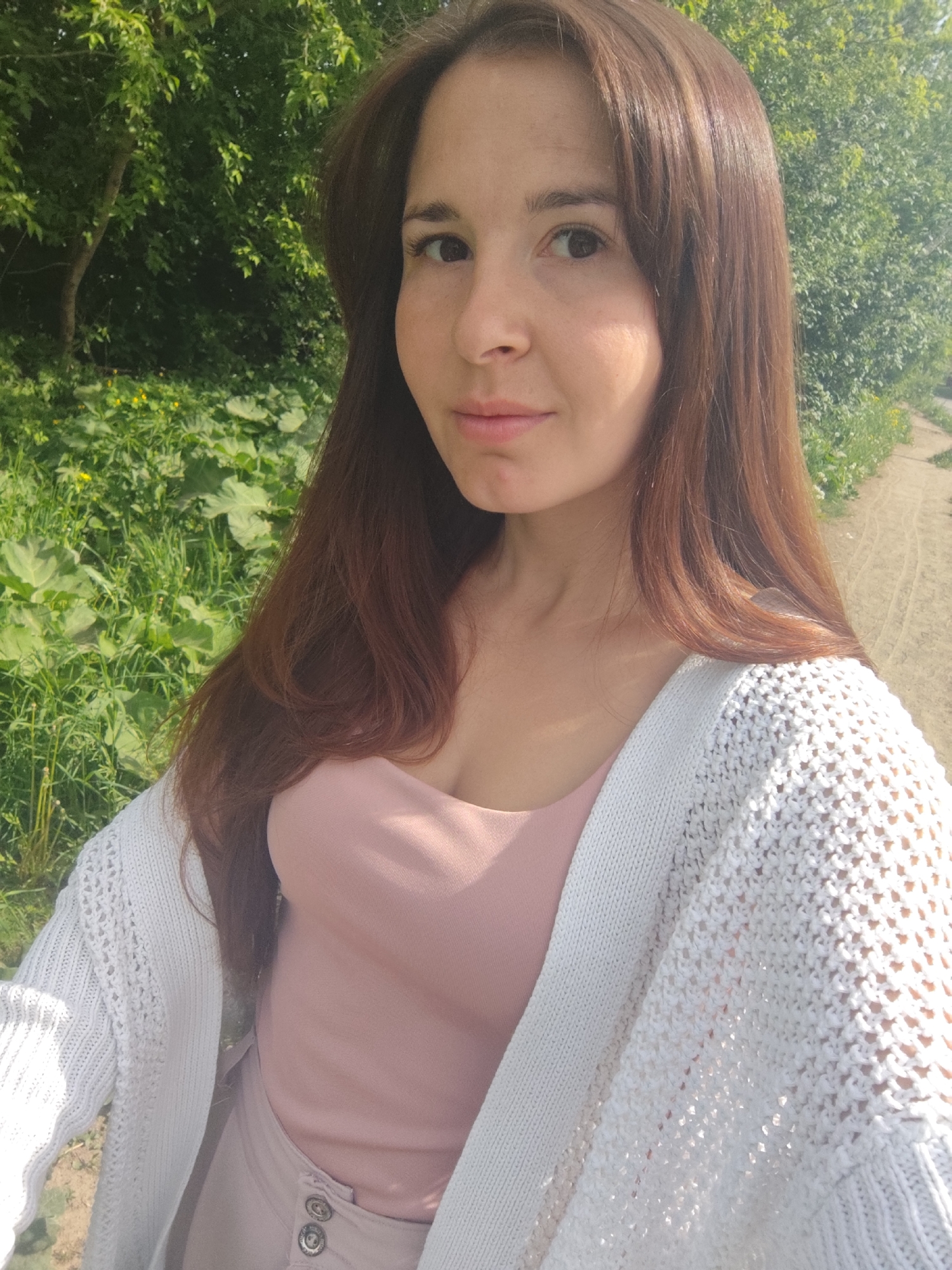 A woman with long brown hair is posing for a selfie in an outdoor environment in Фоки, Russia. She is wearing a pink top and a white knitted cardigan. The background includes green foliage and a dirt path.