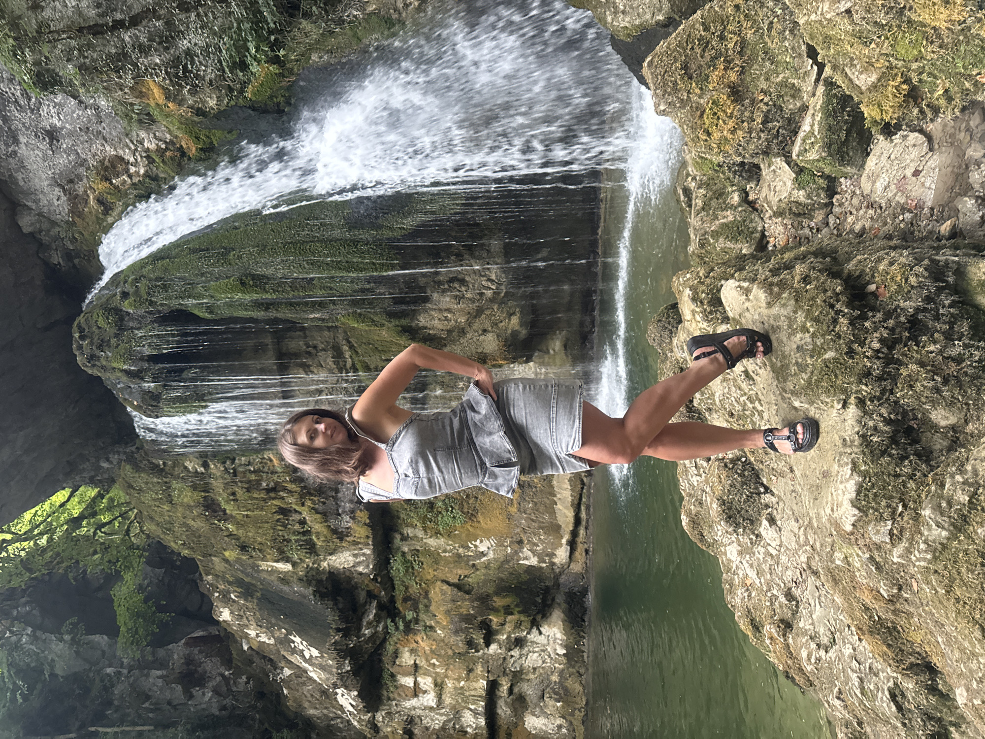 A woman in a gray denim dress and black sandals poses on rocks in front of a waterfall. The waterfall cascades down moss-covered rocks into a pool of green water. The background consists of lush green foliage and rock formations. The image appears to have been taken in a natural outdoor setting, likely a park or nature reserve. Based on the context of Saint-Claude, France, this is likely the Cascade du Flumen.