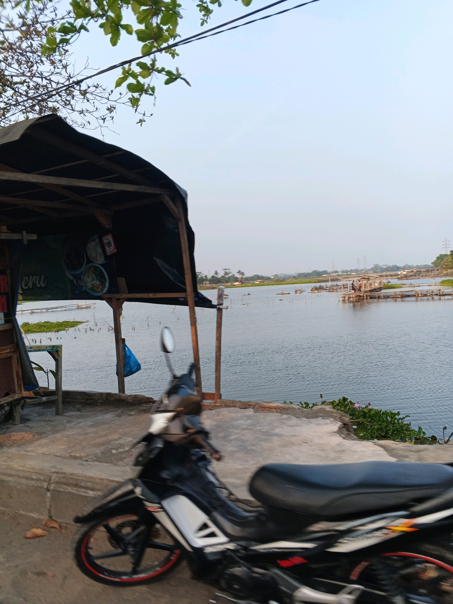 The image depicts a waterfront scene in Cilacap, Indonesia, likely near the mangrove areas or fishing ponds. In the foreground, a black and white motorcycle is parked, with its front wheel slightly blurred due to motion or focus. The motorcycle has red detailing on its rim. Behind the motorcycle, a simple wooden structure with a dark, possibly tarped, roof serves as a small roadside stall or shelter. A menu is visible hanging under the roof, showing pictures of food. The stall is situated on a concrete embankment overlooking a large body of water. The water is calm, with some aquatic plants dotting its surface. In the distance, wooden structures resembling fish farms or small jetties with people can be seen on the water. The sky is a pale blue, indicating either early morning or late afternoon. Power lines stretch across the upper part of the frame, and distant trees and buildings are visible along the horizon. The overall impression is of a rural or semi-rural setting by a body of water, likely a place where local food is sold to travelers or visitors.