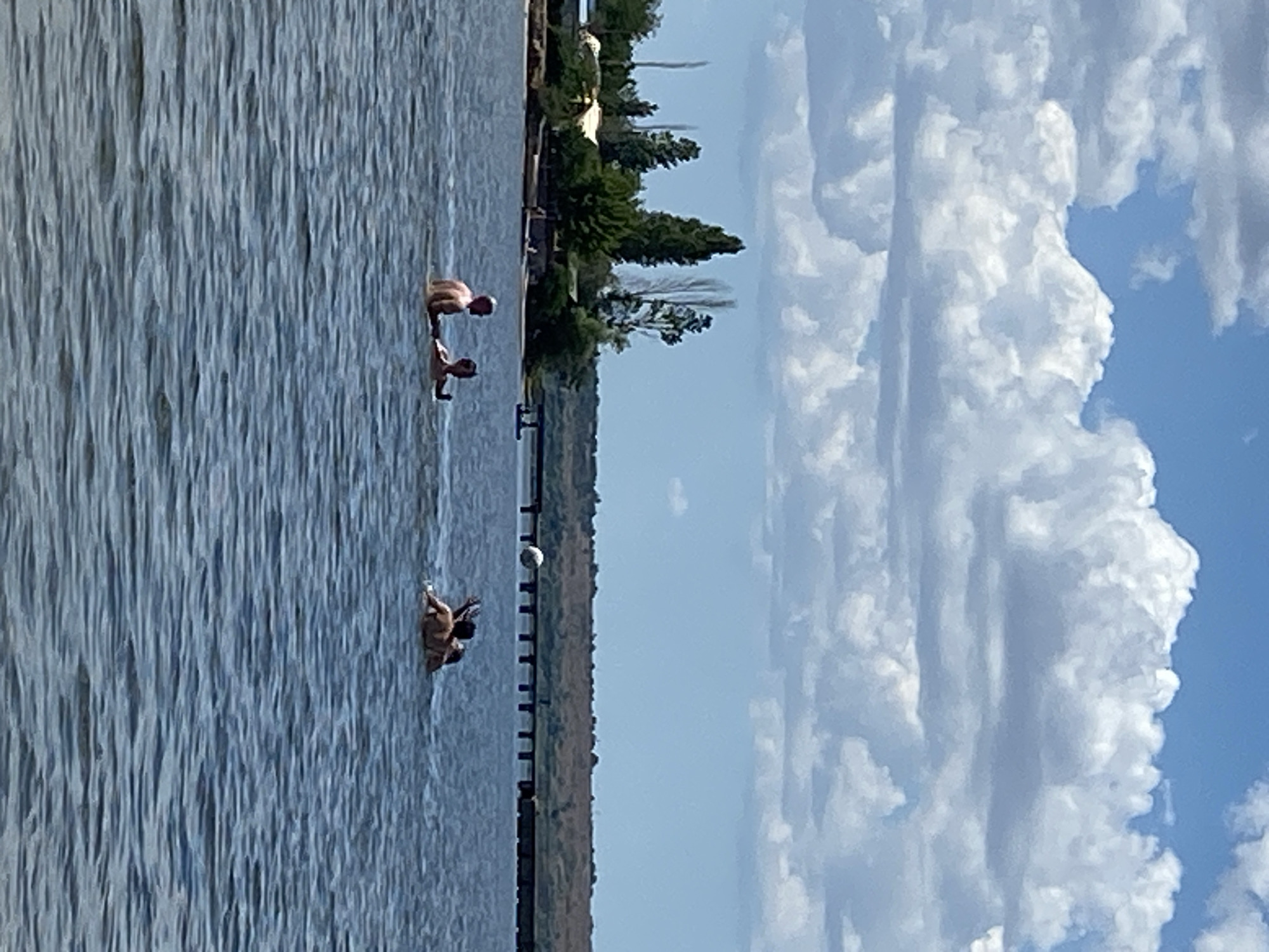A wide shot captures a serene body of water under a sky filled with fluffy white clouds. The water is a deep blue, with gentle ripples distorting the reflections of the sky and distant objects.
In the foreground, several people are partially submerged in the water. Two individuals are closest to the viewer, with one appearing to be talking to the other, both facing left. Further to the right, another group of individuals are engaged in what seems to be a game of volleyball, with a white ball visible in the air.
The middle ground of the image is dominated by the calm water, with a distant shoreline dotted with trees and some structures. A bridge with numerous pilings stretches across the water in the background, connecting the two sides of the body of water. The sky above is a vibrant blue, with large, dramatic cumulus clouds adding texture and depth to the scene. The overall atmosphere is one of leisure and outdoor recreation on a sunny day.