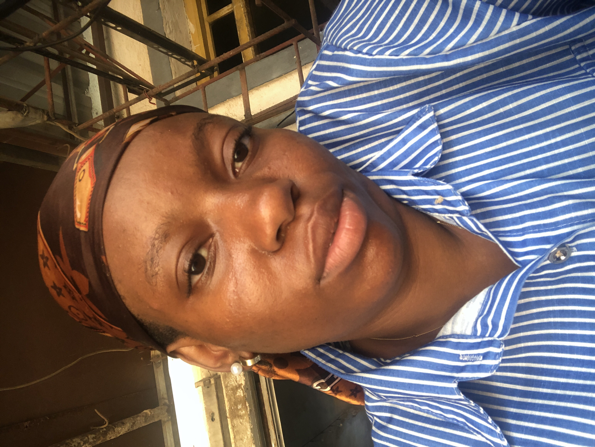 A close-up portrait captures a person, likely a woman, with dark skin, looking directly at the camera. She wears a blue and white striped collared shirt, and a brown headscarf with an indistinct pattern or symbols on it. A white pearl earring is visible on her left ear, and a thin gold chain around her neck. The person has a neutral expression. In the background, there is a metallic structure composed of bars and what appears to be a window or opening, suggesting an outdoor or semi-enclosed setting. The lighting indicates it is daytime. No discernible text is visible on the headscarf or in the background.