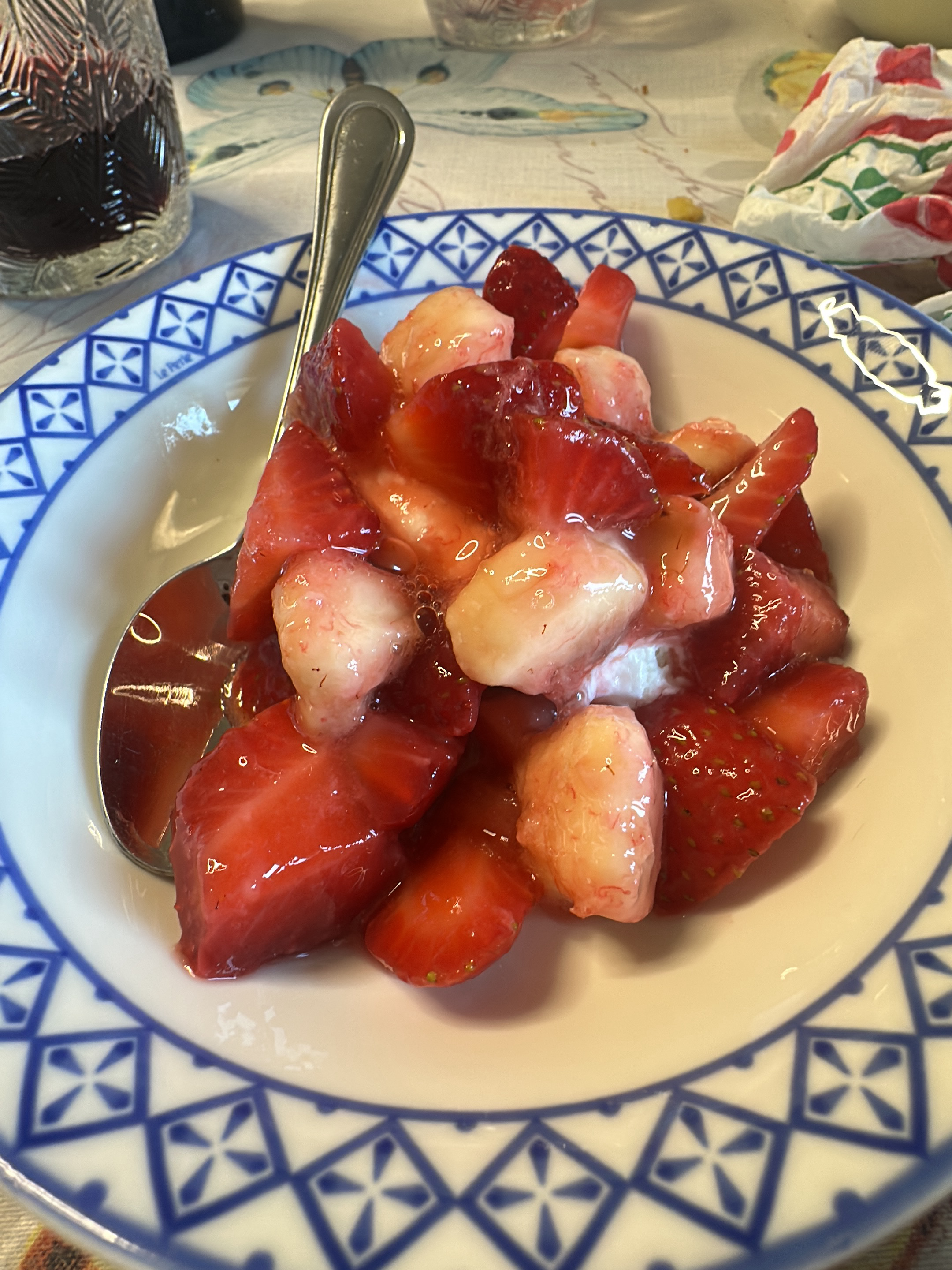 A close-up shows a bowl of sliced strawberries in a clear liquid, likely a glaze or syrup. A silver spoon rests on the left side of the bowl, submerged in the glaze. The bowl itself has a white interior and a blue and white geometric pattern around the rim. The background features various objects and surfaces, including a glass of red liquid, possibly wine, and a patterned tablecloth. The image was taken in Aradeo, Italy.