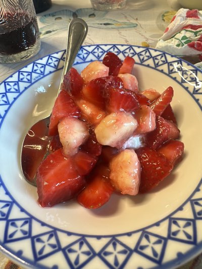 A close-up shows a bowl of sliced strawberries in a clear liquid, likely a glaze or syrup. A silver spoon rests on the left side of the bowl, submerged in the glaze. The bowl itself has a white interior and a blue and white geometric pattern around the rim. The background features various objects and surfaces, including a glass of red liquid, possibly wine, and a patterned tablecloth. The image was taken in Aradeo, Italy.