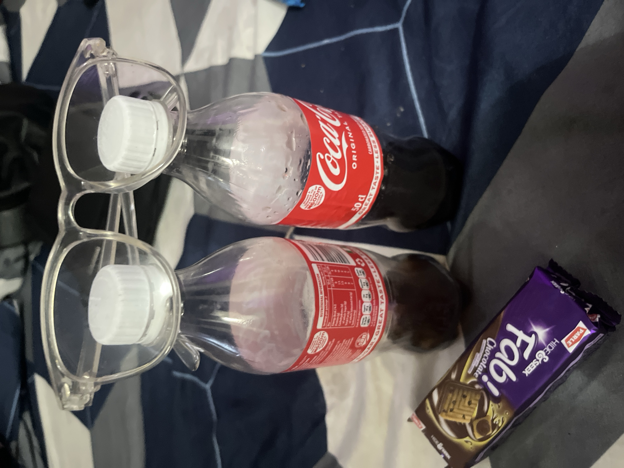 The image shows a scene with several items laid out on a surface with a dark blue and white geometric pattern. Two clear plastic glasses frames are positioned over the necks of two clear Coca-Cola bottles, each containing a small amount of liquid. Next to the bottles is a purple "Fab!" Hide & Seek chocolate cookie bar. The presence of a Coca-Cola bottle labeled "50 cl" and with text stating "GREAT TASTE LESS SUGAR" suggests the image was taken in a region where Coca-Cola products are available. There is no explicit evidence in the image to pinpoint the location as Lokoja, Nigeria. No people or actions are visible.
