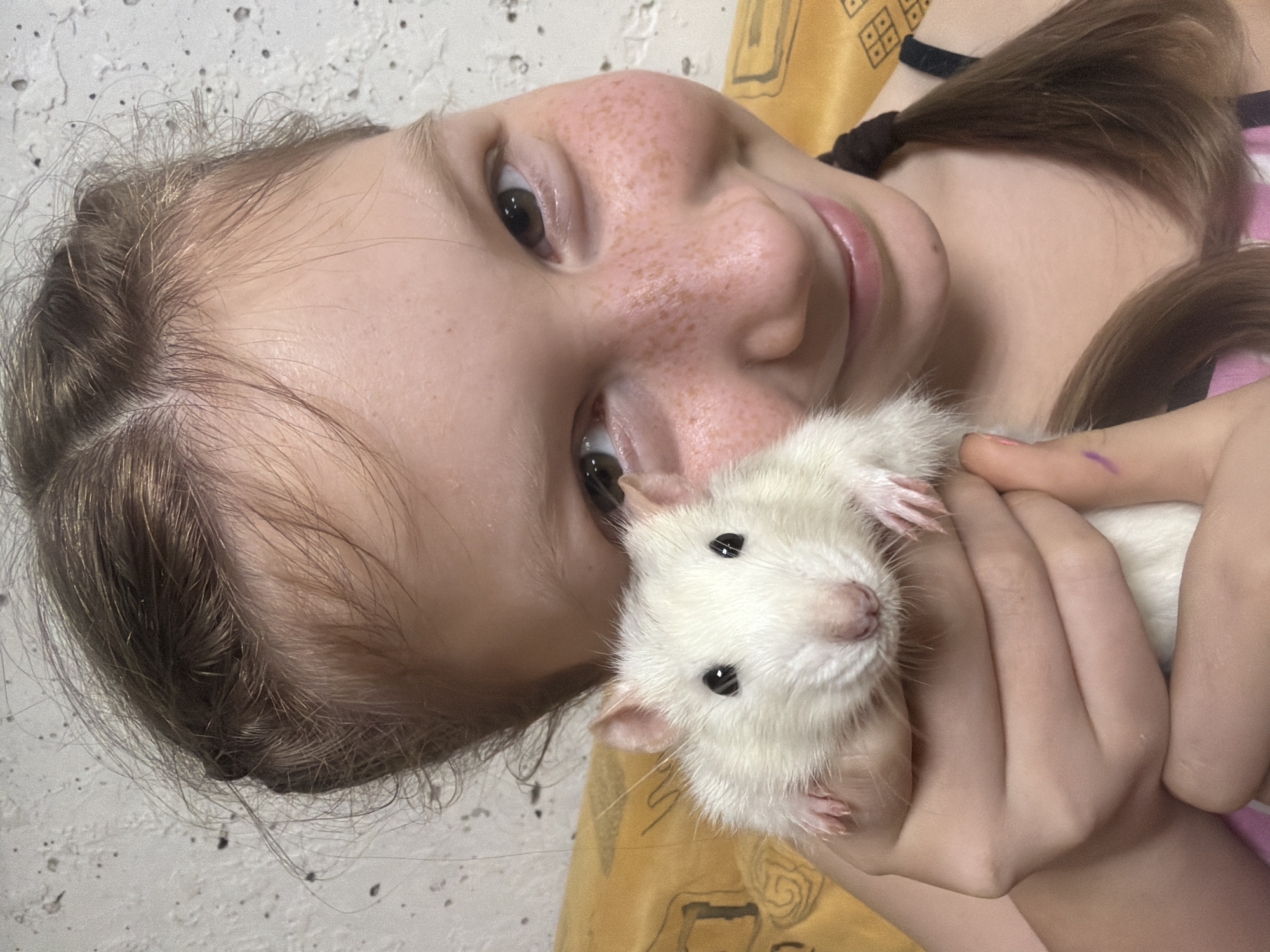 A young girl with freckles is holding a white rat. The girl is smiling and looking at the camera. The rat is also looking at the camera. The background is a light gray wall and an orange and yellow patterned fabric. It is not possible to verify the city of Markovo, Russia.