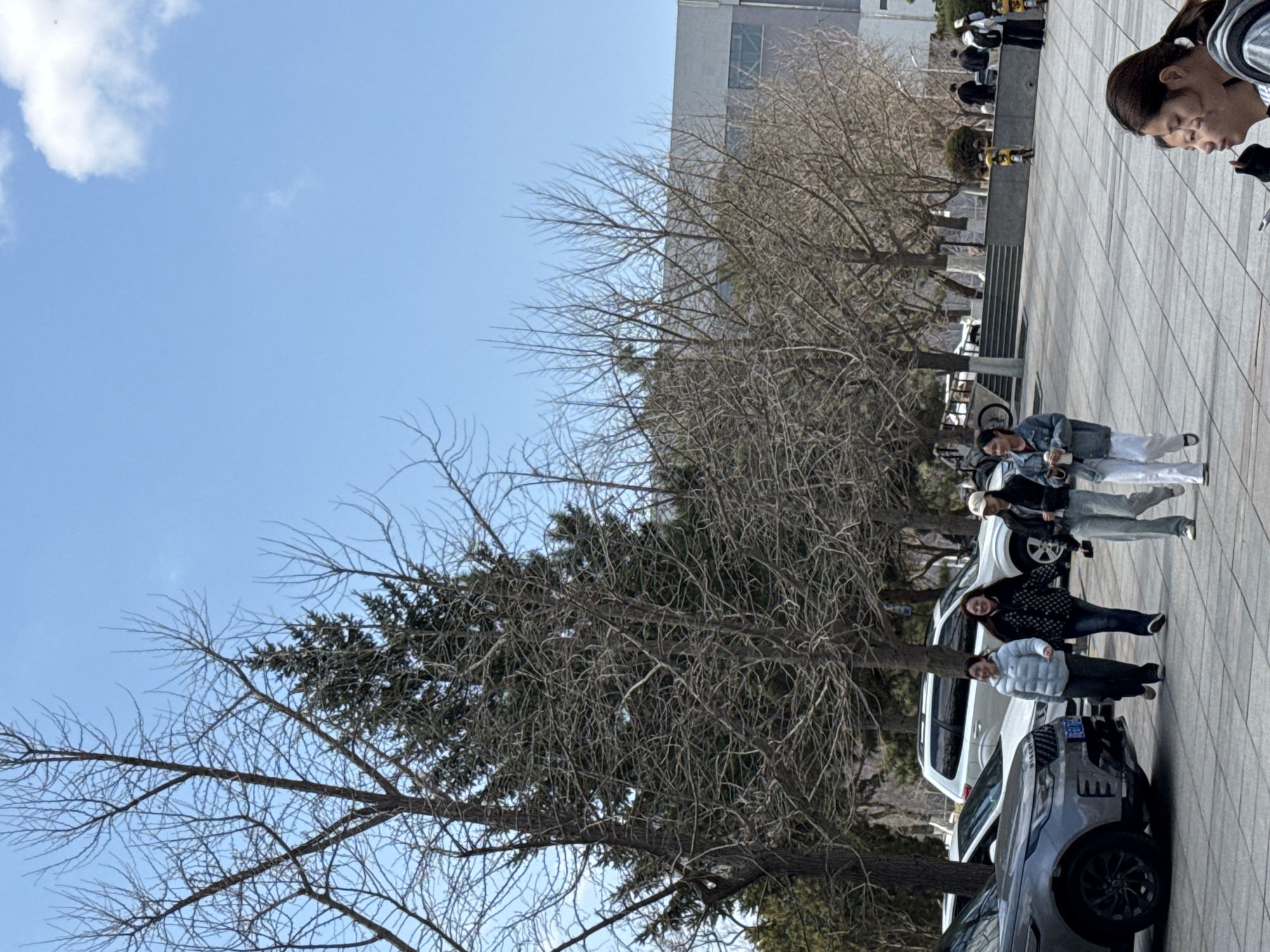 A daytime exterior shot shows people and vehicles on a paved surface adjacent to trees, a building, and an elevated platform with people. The sky is blue with some clouds. A gray SUV with a black wheel is parked on the right edge of the image. Next to it are white vehicles. Four people are walking, with one person visible in the top right corner. On the elevated platform are people in uniform. The location is likely Zhongtun, China, based on the prompt.