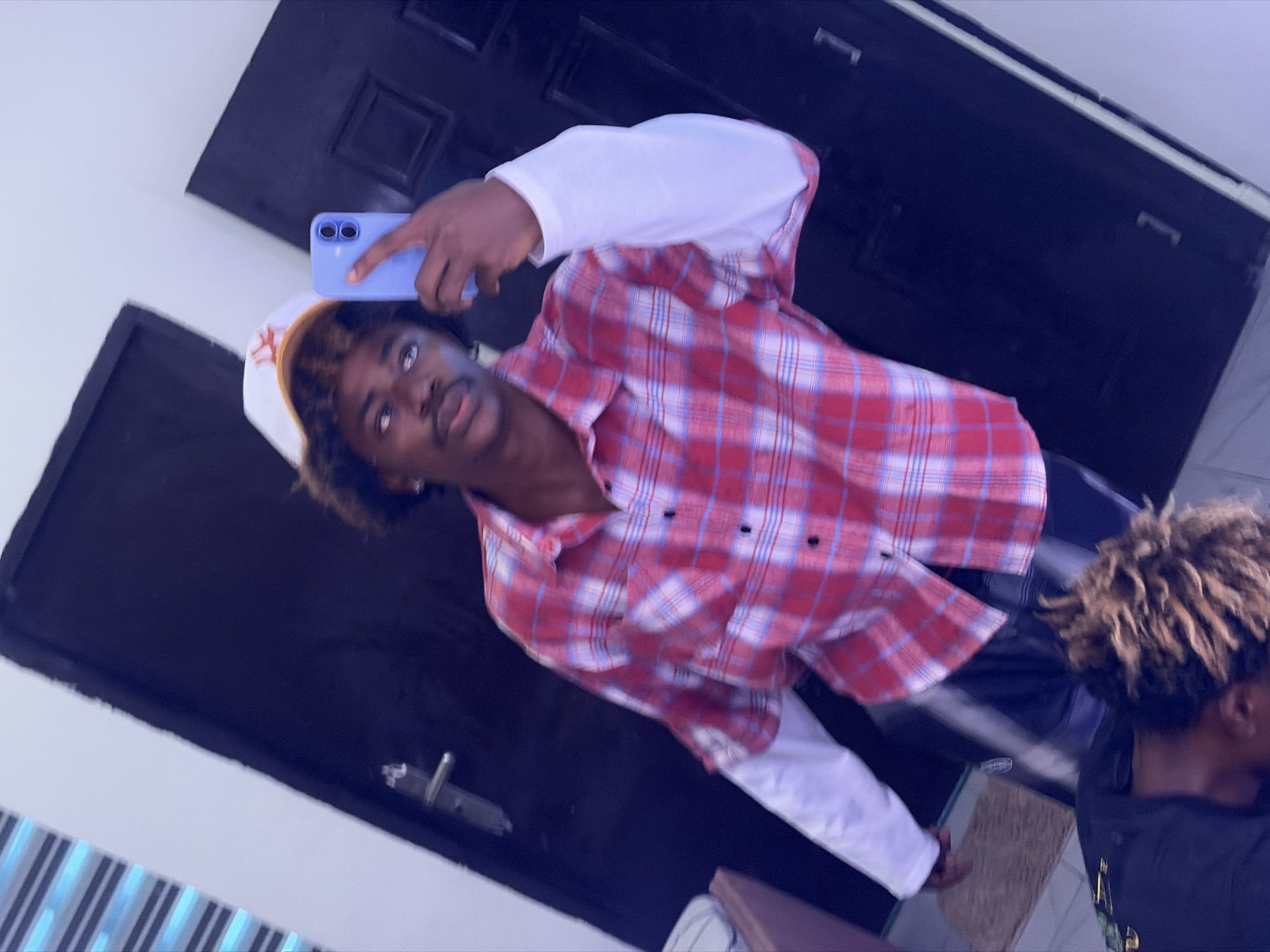 The image, captured in Igboefon, Nigeria, presents a tilted, casual indoor scene, likely a selfie. The main subject, a young person with dark skin and dark, curly hair, is positioned in the center, looking directly into the camera lens of a light blue smartphone held up. They wear a white baseball cap with an orange design on the front, a red and white plaid button-up shirt unbuttoned over a white long-sleeved shirt, and dark pants. Their expression is neutral with slightly parted lips.
The background consists of dark, possibly double, doors on the left and right, with light-colored walls above and between them. The floor features a striped pattern to the far left and a small, light-brown rectangular mat-like object on the right. A second individual is partially visible on the far right, with only the top of their head showing light brown dreadlocks and a dark shirt, mostly out of focus. The overall lighting suggests an indoor setting during daytime. No discernible text is visible on clothing or objects.