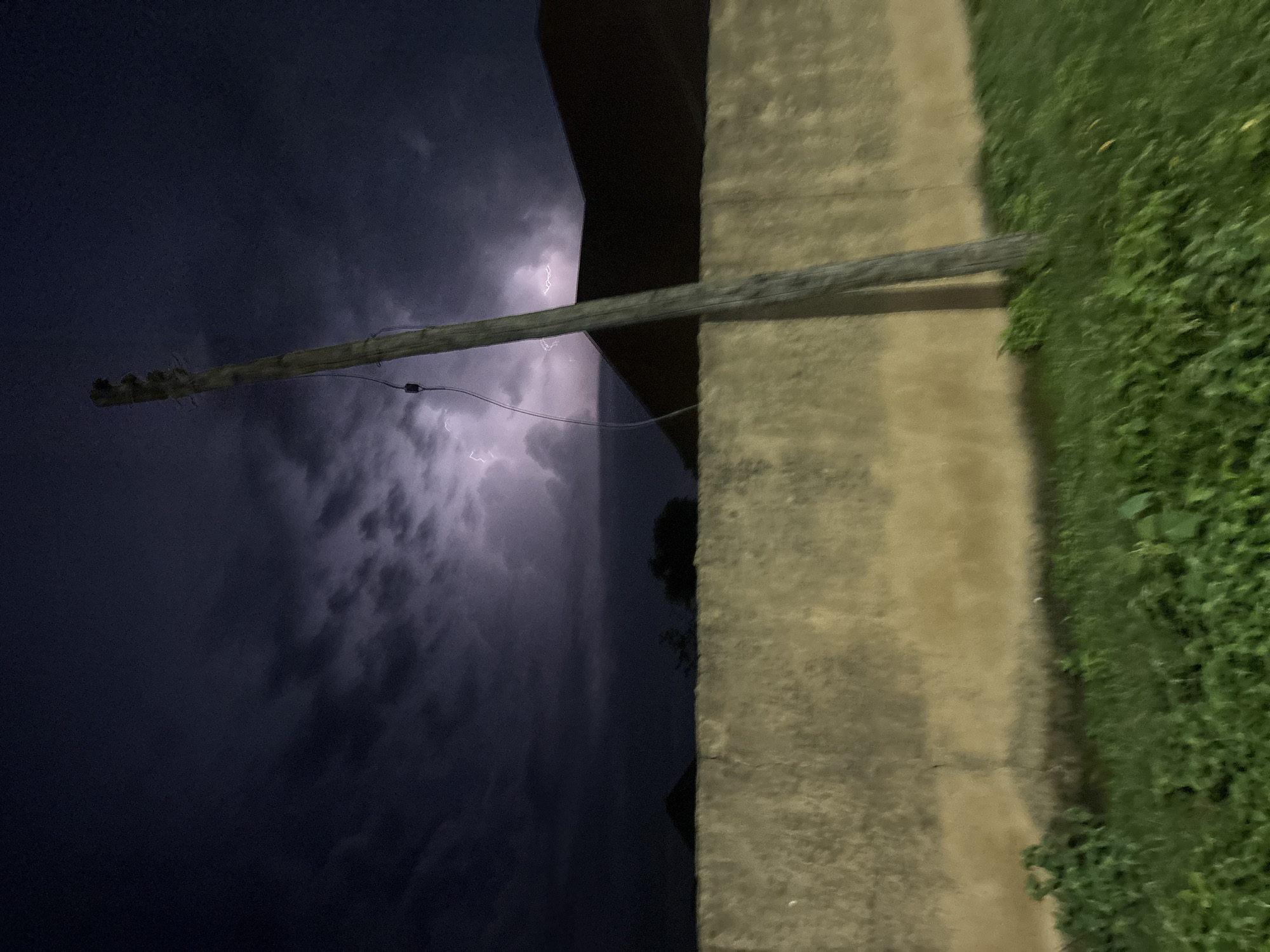 This image captures a nighttime scene in Bukuru, Nigeria, featuring a dramatic thunderstorm. A tall, weathered utility pole stands prominently in the foreground, leaning slightly to the right and bisecting the frame. A wire is visible running down the pole. Behind the pole, a thick, textured concrete wall stretches horizontally across the mid-ground. Beyond the wall, the dark silhouettes of buildings, possibly houses with gabled roofs, are visible against the stormy sky. The sky itself is a dark, turbulent mass of clouds illuminated by flashes of lightning, creating streaks of bright light in the upper portion of the image. The foreground is covered in lush green grass, some of which appears slightly out of focus. The overall impression is one of a powerful natural event occurring in a residential or semi-urban setting.