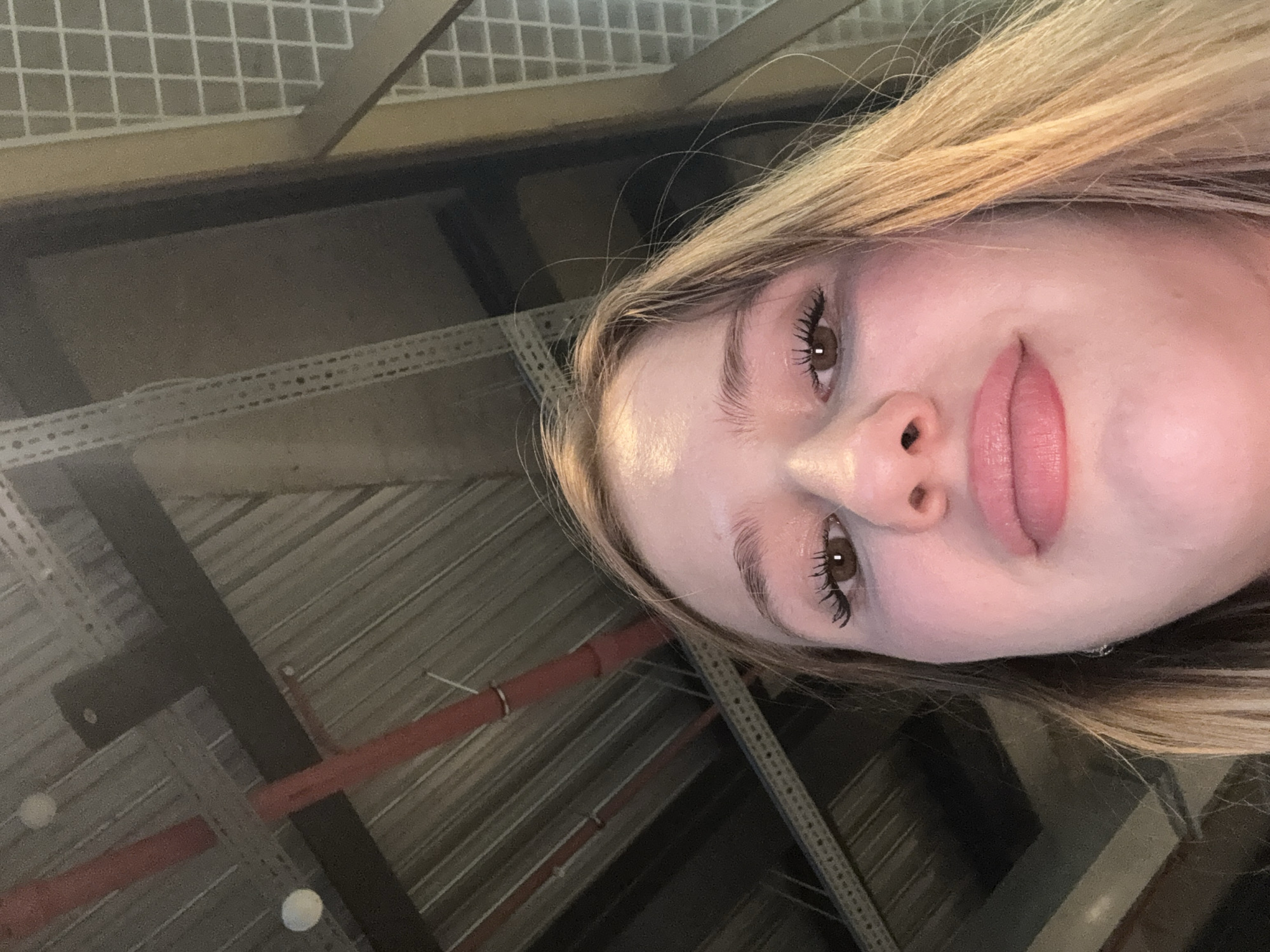 The scene is located in Sinyavino, Russia. The image displays a low-angle, close-up view of a young woman's face, positioned centrally and looking directly at the camera. Her brown eyes are open, featuring distinct long eyelashes. Her lips are full and bear a light pink hue. Light brown hair, possibly with blonde highlights, frames the right side of her face. The background consists of an industrial-style ceiling, characterized by corrugated metal decking, prominent dark structural beams, and two red pipes running diagonally across the frame. Two small, circular white lights are visible on the ceiling. On the far right, a vertical structure with a white grid pattern is partially visible.