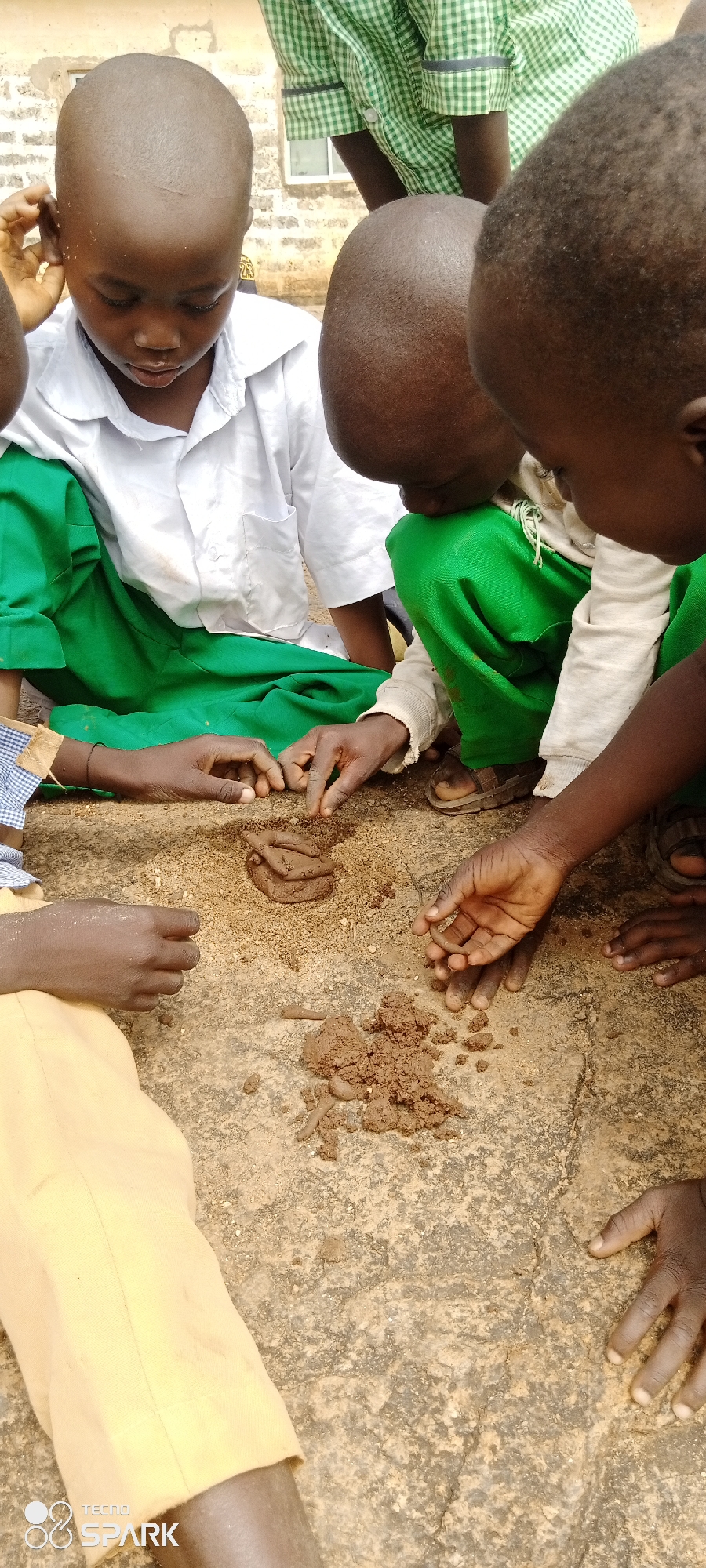 In Kagarko North, Nigeria, a group of young children are engaged in an activity with mud on a rough, outdoor surface. The children are wearing school uniforms, with one child in a white shirt and green skirt, and another in green trousers. Their hands are actively manipulating the mud, which is formed into small piles and sticks on the ground. The environment appears to be a schoolyard or similar outdoor area, with a weathered wall in the background. The children's focus is on their shared task, with some peering intently at the mud.