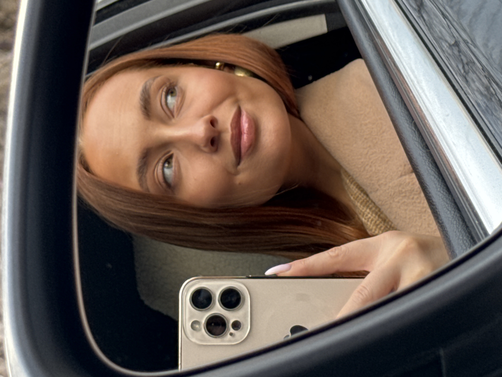 The image is a reflection of a woman taking a selfie with her smartphone in what appears to be a car's side mirror. The woman has light skin, reddish-brown hair, and is wearing gold hoop earrings. She is wearing a light-colored coat. The smartphone is gold. The setting seems to be inside a car, with parts of the interior and exterior visible in the reflection. There's no information to confirm the location is Chaikovsky, Russia.