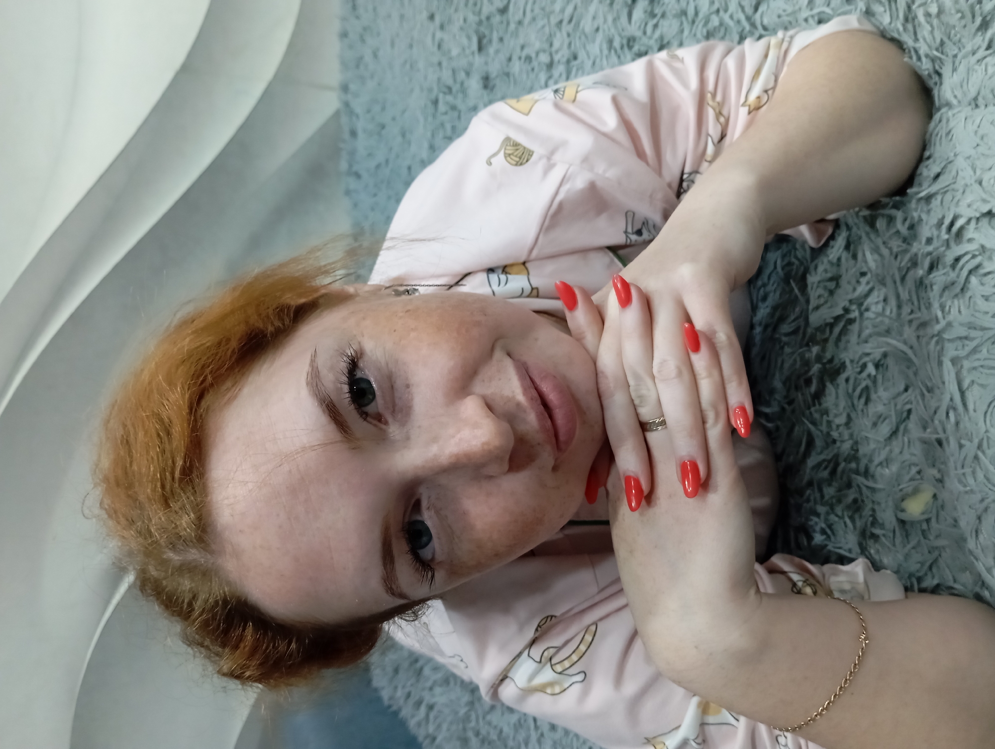 A woman with freckles and red hair is lying on a grey rug, looking at the camera. She is wearing a pink pajama top with illustrations of bananas and cats. Her nails are painted red, and she is wearing a gold ring on her finger and a gold bracelet on her wrist. The location is presumably indoors. The image suggests it was taken in Yurgamysh, Russia.