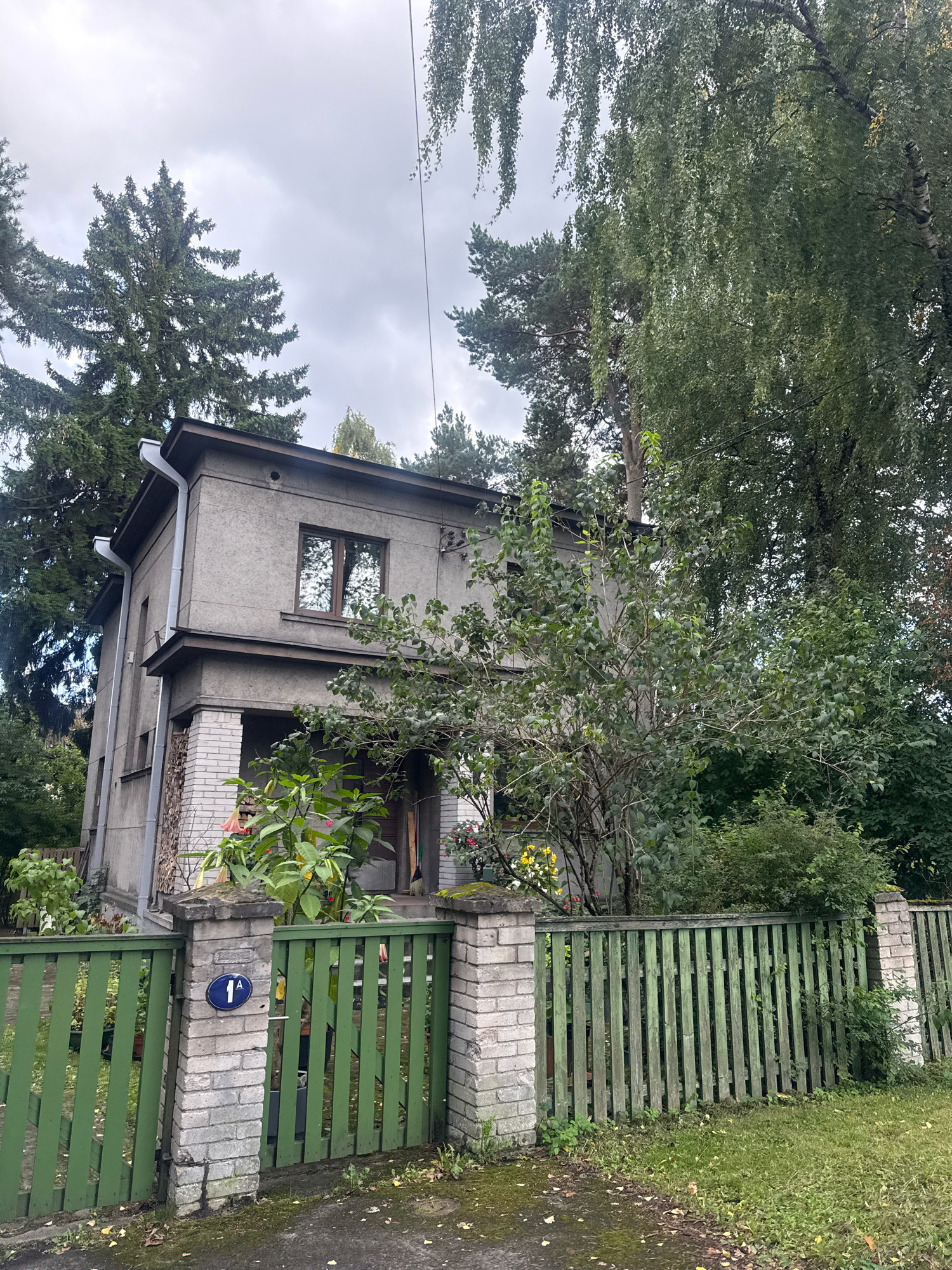 The image shows a two-story, grey-rendered house with a dark brown roof and window frames, located in Tallinn, Estonia. The house has a porch with brick pillars. In front of the house, there is a green wooden fence with brick pillars. A blue oval sign with the number "1A" in white is affixed to one of the pillars. Mature trees, including a pine and a birch, surround the property. The sky is overcast with grey clouds.