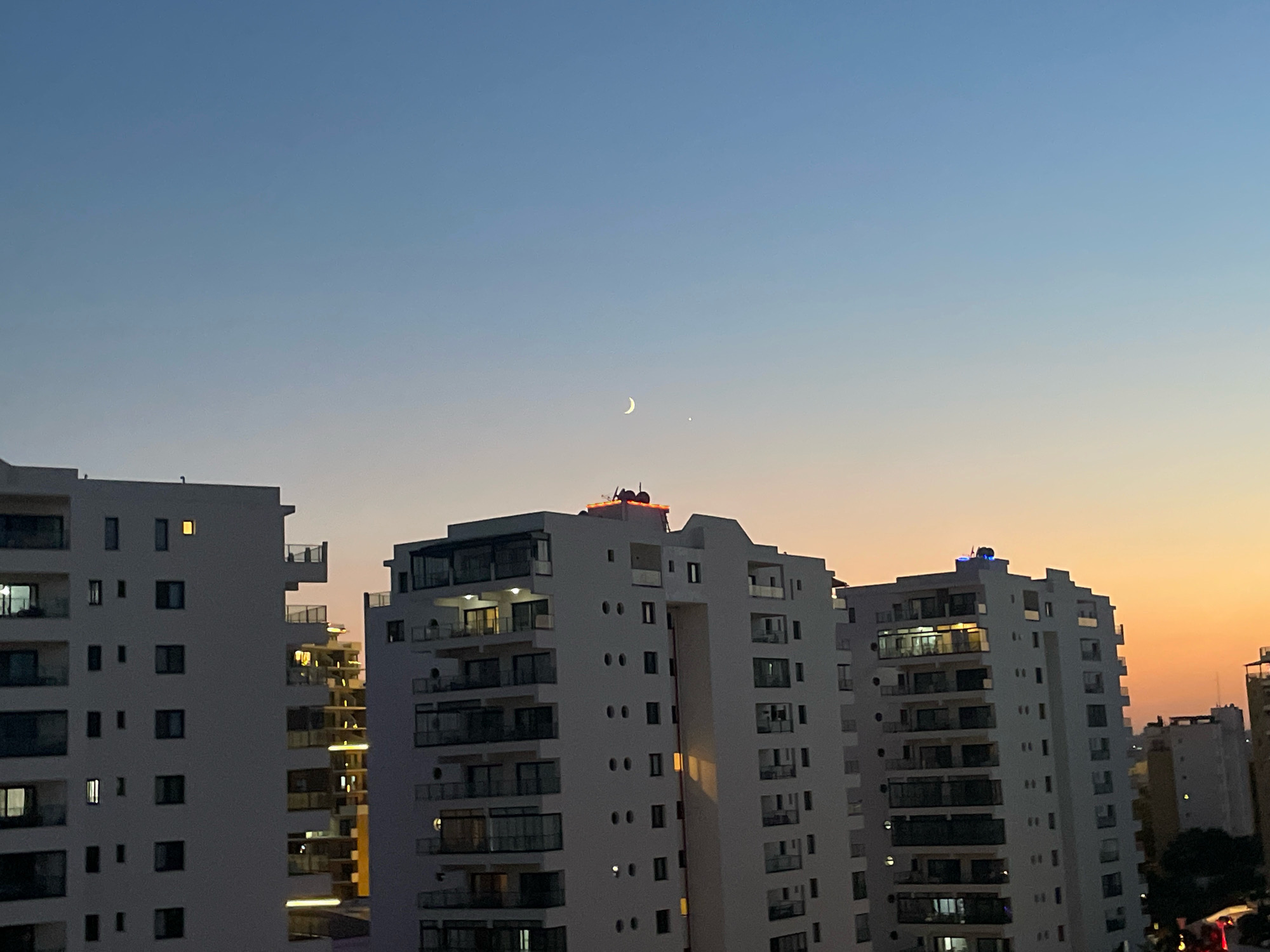 The image depicts a dusk sky over the city of Trikomo, Northern Cyprus. The sky transitions from a pale blue at the top to a warm orange hue near the horizon, illuminated by the setting sun. A crescent moon and a bright star are visible in the upper portion of the sky. In the foreground, several modern multi-story apartment buildings stand tall, their white facades contrasting with the darkening sky. Some windows are lit from within, indicating occupancy. The architecture of the buildings appears contemporary with balconies and numerous windows. The overall scene captures a peaceful urban twilight in Cyprus.