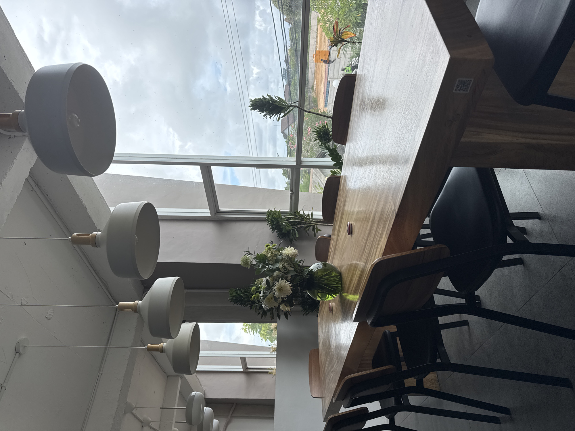 The image shows the interior of a space, possibly a cafe or restaurant, with a large wooden table as the main focus. The table is set with chairs and has decorative plants in vases. A row of pendant lights hangs from the ceiling, adding to the ambiance. The scene is well-lit, with natural light coming through large windows that offer a view of an outdoor landscape. The location is Unknown, Unknown.