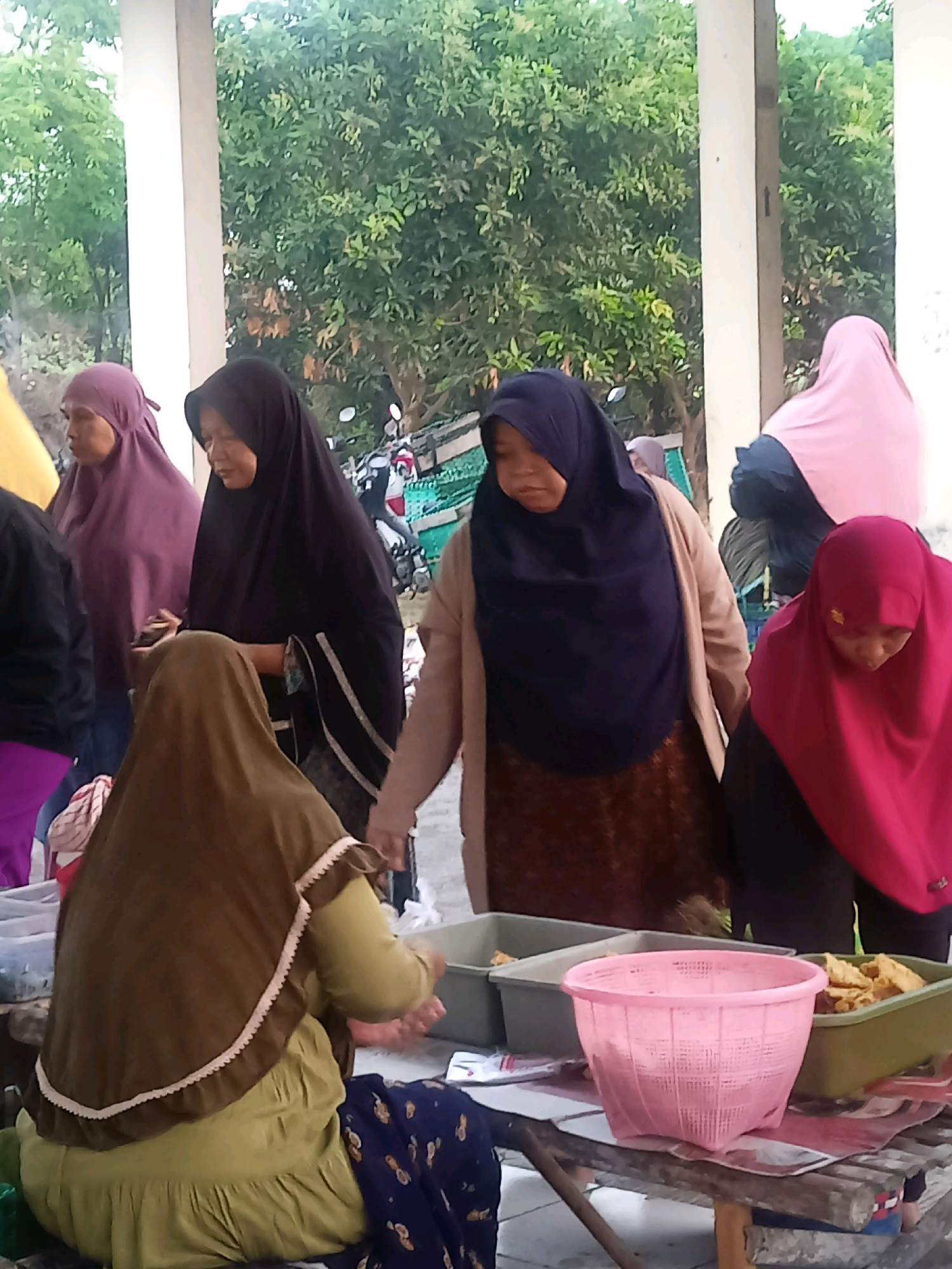 In Nganti, Indonesia, a group of women, some wearing hijabs, are gathered around tables outdoors. In the foreground, a woman in a brown hijab and a light green dress is seated, facing away from the camera. To her right, a pink plastic basket sits on a wooden table laden with what appear to be fried food items. Behind her, other women are visible, some interacting with each other. One woman in a black hijab and a tan cardigan is looking down, while another in a dark blue hijab and a brown patterned skirt is also looking downwards. Further back, a woman in a pink hijab is partially visible, and a woman in a purple hijab is standing. The background consists of trees and the weathered pillars of an outdoor structure. The overall scene suggests a casual gathering, possibly a market or a communal event, with food being a central element.
