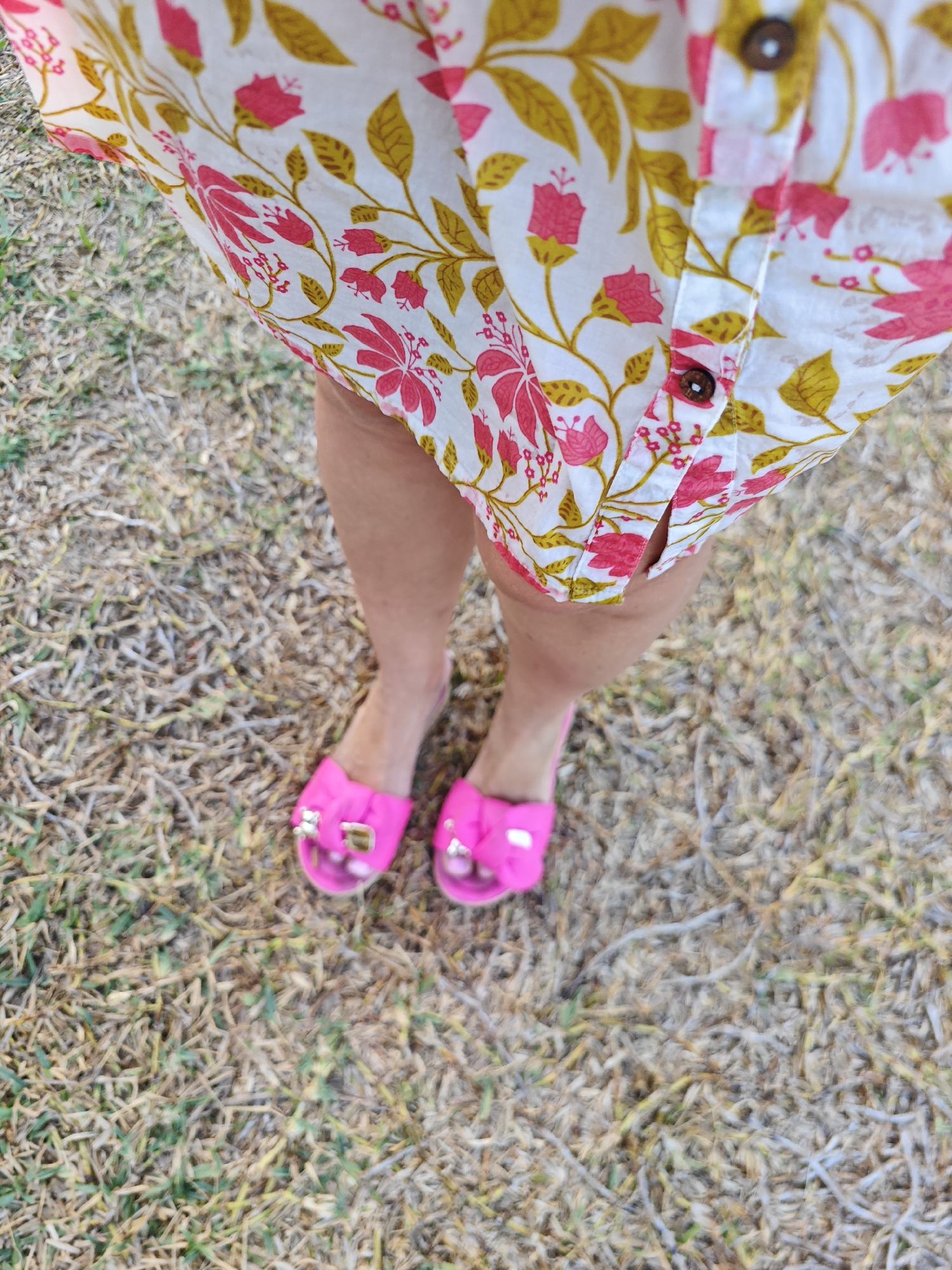 A close-up, high-angle shot shows a person's legs and feet from the knees down. They are wearing a white shirt with a floral pattern in pink and yellow, and bright pink slide sandals with gold buckles. The person is standing on dry grass and dirt, which appears to be in an outdoor urban setting. The background is blurred, with hints of dry vegetation and possibly some gravelly ground. The lighting suggests it is daytime. The image has a slightly washed-out appearance. The location is the city of Mahdia, Tunisia.