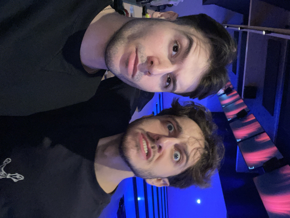 Two dark-haired men pose closely for a selfie. The man on top has dark, smooth hair and a serious expression. Below him, the other man has wavy hair and a surprised, slightly silly expression, baring his teeth. Both wear black shirts. The background is a mix of blue and dark tones, with a structure featuring horizontal bars displaying a pink and black pattern. The photo appears to be taken indoors, possibly in a club or entertainment venue.