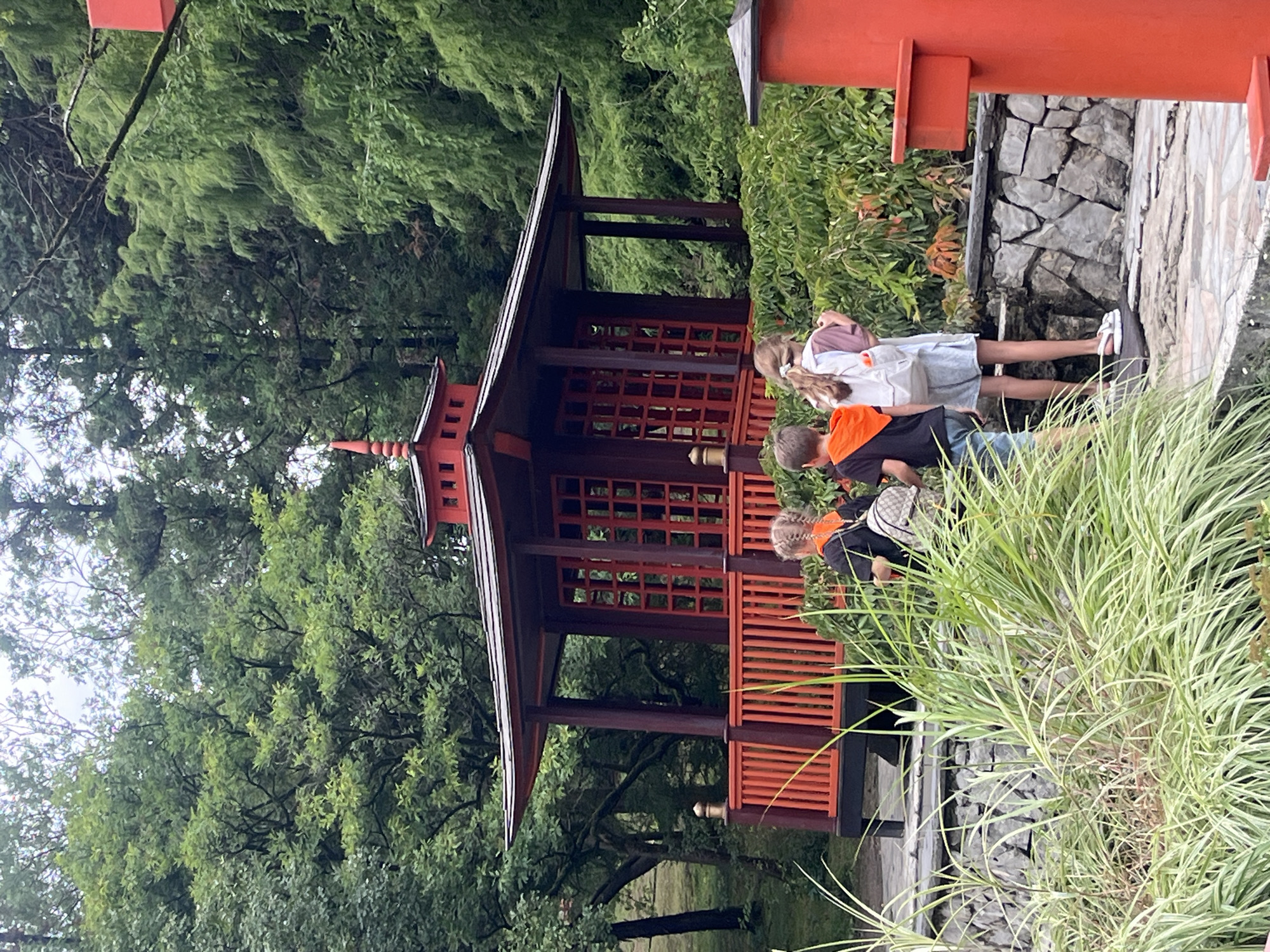Three children, two boys and one girl, stand in a park with a Japanese-style pavilion. The boy on the left wears a black t-shirt with an orange hood and blue shorts. The boy in the middle wears a black t-shirt with an orange hood and blue shorts. The girl on the right wears a light purple t-shirt, denim shorts, and a white backpack. They are all facing away from the camera, looking at the pavilion. The pavilion is red with intricate wooden lattice work. The park has lush green trees and foliage, and a stone wall. In the foreground, tall green grass grows. A red decorative post is visible on the right side of the image.