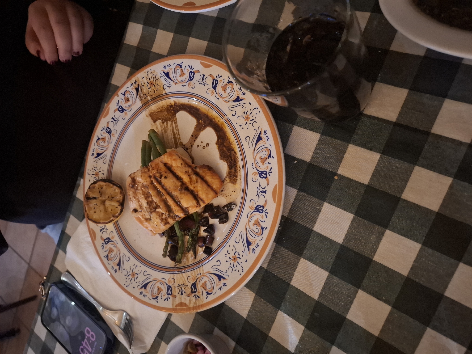 The image shows a plate of grilled salmon with green beans, lemon, and black olives on a patterned plate. A glass of dark liquid, possibly wine, sits to the right of the plate. A person's hand is visible to the left. The plate rests on a green and white checkered tablecloth. A cell phone and fork are visible below the plate. The location is possibly in Naucalpan de Juárez, Mexico, but this cannot be determined definitively from the image alone.