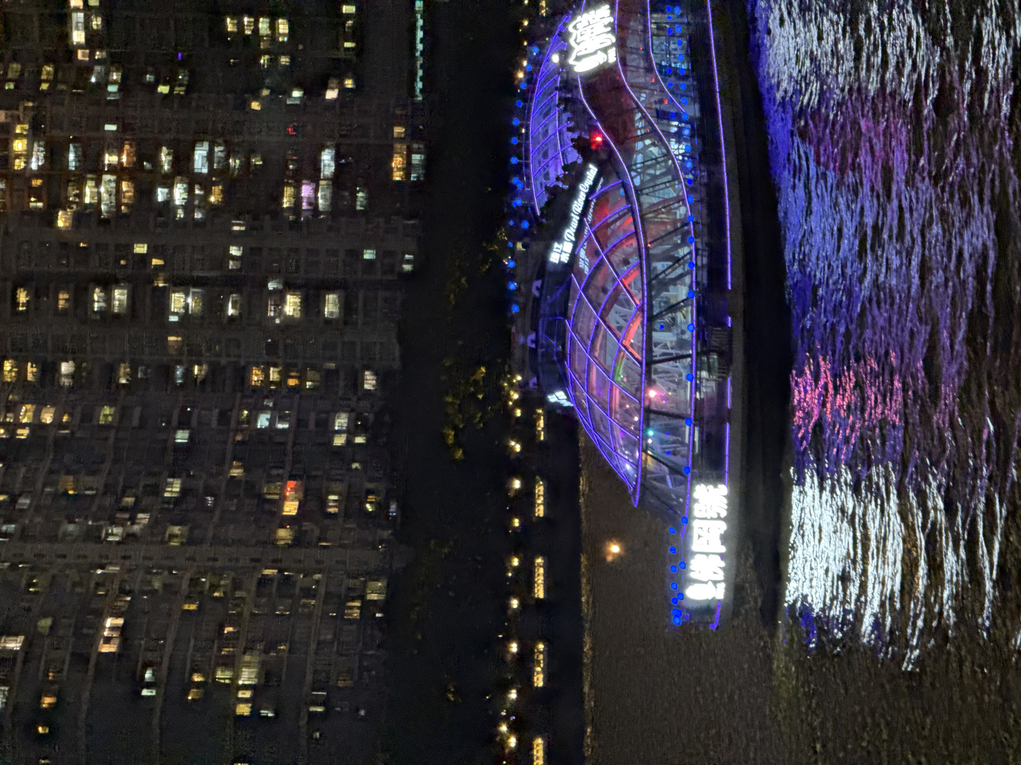The image depicts an nighttime cityscape in Yuexiu District, China, featuring buildings with illuminated windows on the left, a river with light reflections on the right, and a modern glass building structure with blue and red lighting in the center. The glass building has signage in Chinese and English ("Pearl River Crystal"). The river reflects the building's lights, as well as other white and purple lights. Small lights lining the river are visible on the right side of the river.