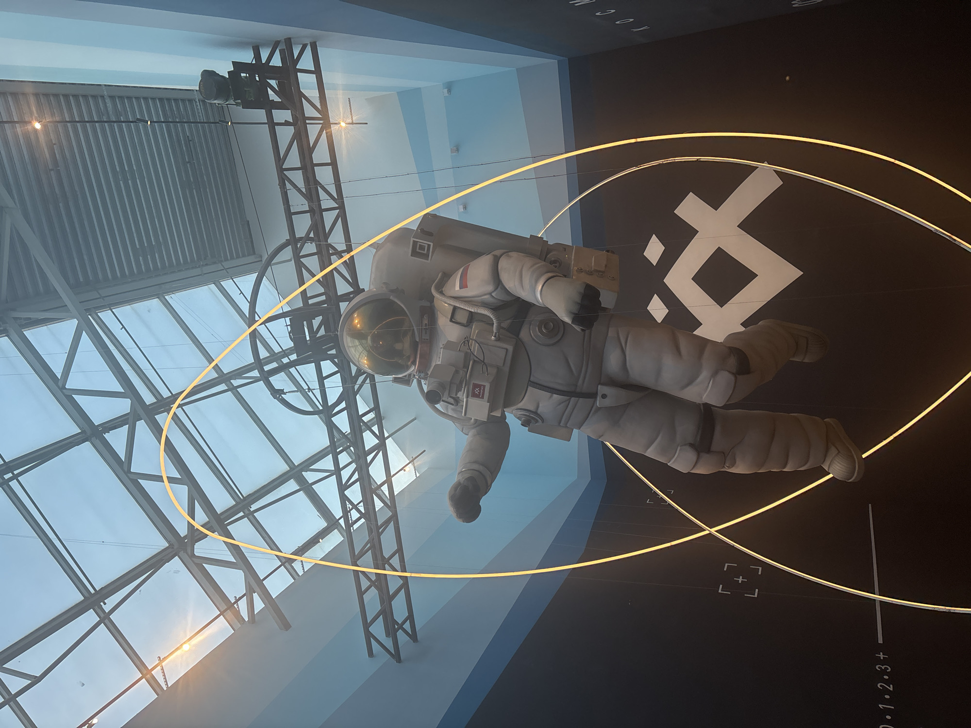 An illuminated sculpture of an astronaut is suspended inside a modern building with a glass ceiling. The astronaut is depicted in a full white spacesuit with a gold-tinted helmet, floating in a dynamic pose. Yellow neon light rings encircle the astronaut, creating an ethereal glow. The background features a black wall with a large white logo and some faint markings resembling a measurement scale. The ceiling is a complex network of metal beams and glass panels, allowing natural light to filter in. The overall impression is that of a futuristic exhibition or art installation, possibly related to space exploration, located within a contemporary architectural space in Saint Petersburg, Russia.