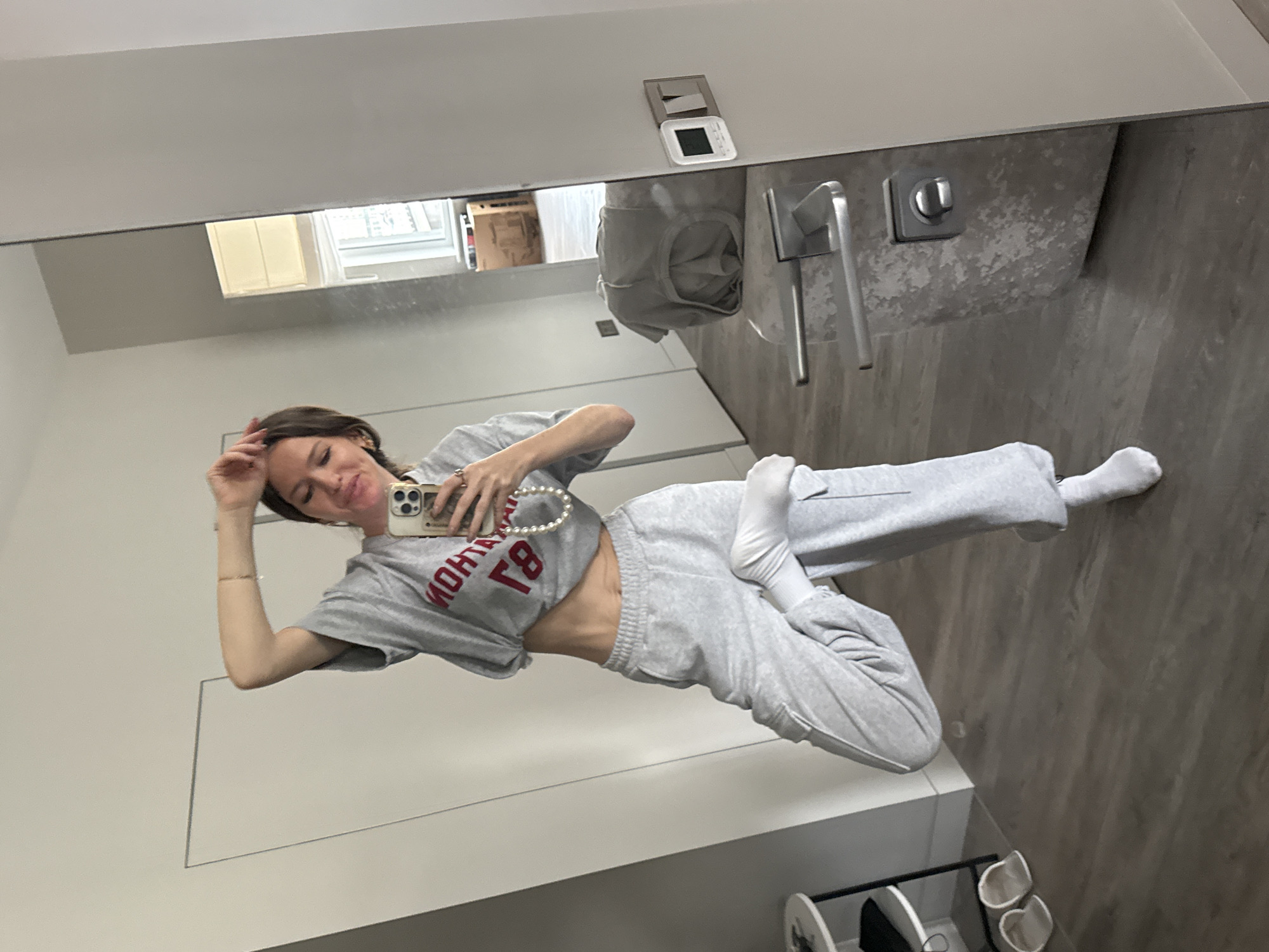 A woman is posing for a selfie in a bathroom mirror. She is wearing a grey cropped t-shirt with red lettering, grey sweatpants, and white socks. She is holding a smartphone with a pearl necklace draped over it. The bathroom has light grey walls and wood-look flooring. There is a towel rack with a grey towel, a light switch and a thermostat on the wall above the towel rack. A shoe rack is visible in the lower right corner. The location is in the city of Unknown, Unknown.