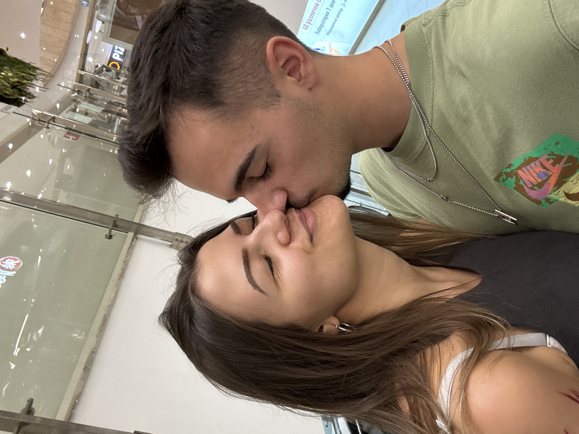The image portrays an intimate moment between a man and a woman in an indoor public setting, likely a shopping mall in Sofia, Bulgaria. The man, with dark hair and light stubble, leans in to affectionately kiss the woman on her nose or forehead. He is wearing a light olive-green t-shirt featuring a colorful Nike logo graphic and two silver chain necklaces. The woman, with long brown hair and silver hoop earrings, has her eyes closed, exhibiting a serene and content expression as she receives the kiss. She wears a dark, possibly sleeveless, top.
The background shows reflections from glass panels and bright overhead lighting, characteristic of a well-lit interior during the day. Architectural details include glass railings and commercial signage. A partially visible sign reads "PIZ", suggesting a pizza establishment. More distinct Cyrillic text on another sign includes phrases such as "40 различни с" (40 different with...), "Лаборатории 7 дни" (Laboratories 7 days), and "Медицински център" (Medical center), indicating a medical or service facility. The photograph is taken from a tilted angle, adding a dynamic quality to the scene of tenderness and closeness between the couple.