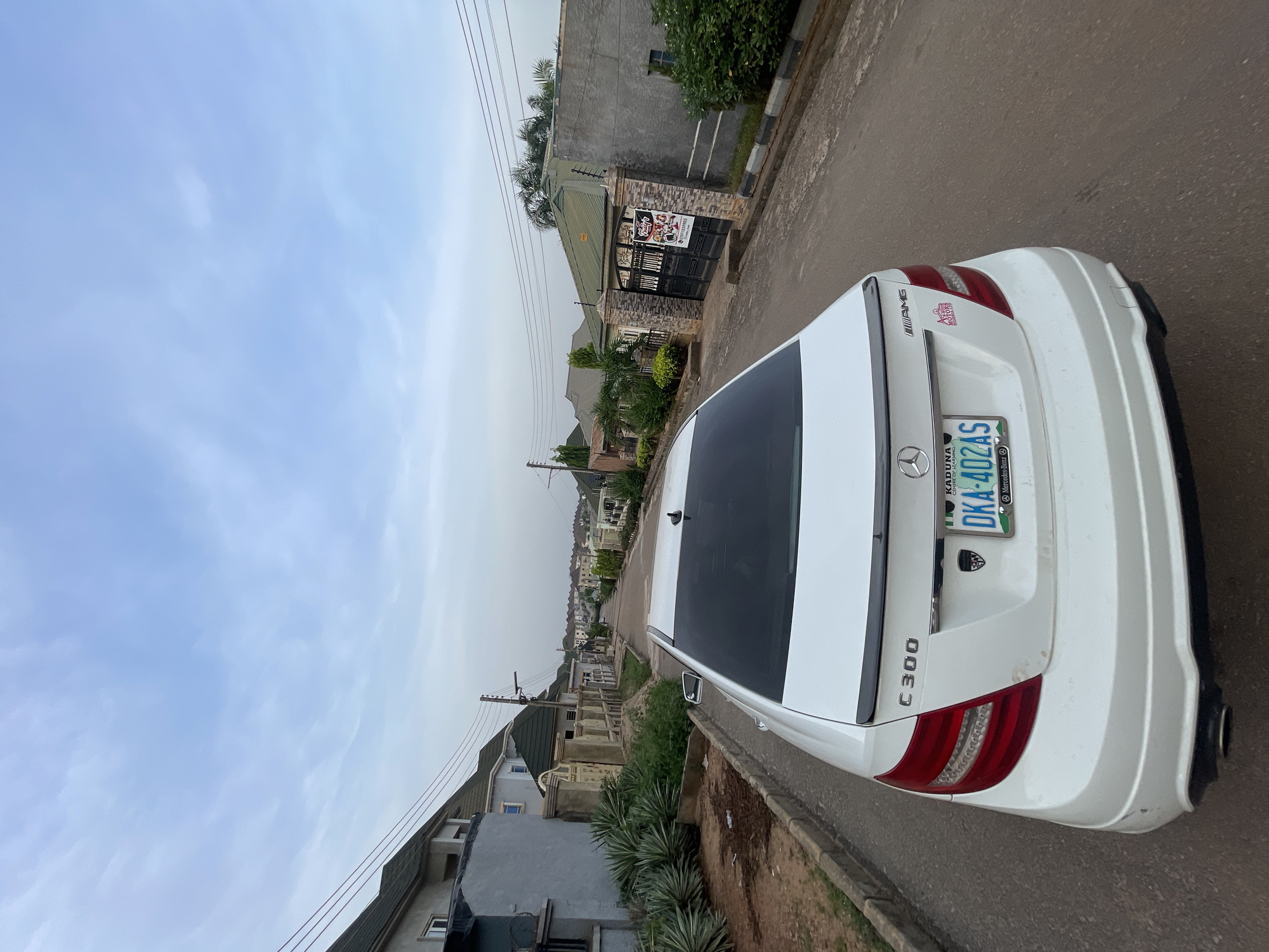 The image displays a white Mercedes-Benz C300 sedan parked or slowly moving on a paved residential street in Abuja, Nigeria, viewed from a slightly elevated angle behind the vehicle. The car's rear is prominently featured, showing the "C 300" model designation on the left and an "AMG" badge on the trunk lid. A Nigerian license plate is visible, bearing "KADUNA State of Origin" and the alphanumeric sequence "DKA-402AS," with a small "Mercedes-Benz" logo below it. A red and white sticker on the trunk lid partially reads "ROYAL MOTORS LTD". The car has visible tail lights and a single exhaust pipe on the right. The white bumper shows some minor dirt or scuffs.
The setting is a residential area with several houses lining the street on both sides. The architecture includes concrete block walls, various roof styles, and decorative gates. One prominent gate on the left features a banner with text including "PASTRIES," "FOOD," and "DRINKS," along with a phone number starting "0808". Lush green vegetation, including palm trees and other tropical plants, is present around the houses and along the roadside. Overhead power lines are also visible.
The sky is overcast or hazy, indicating daytime but not direct bright sunlight. No people are visible in the frame, and there is no specific activity or event taking place other than the presence of the car on the street. The overall impression is a quiet street scene in an urban or suburban area.
