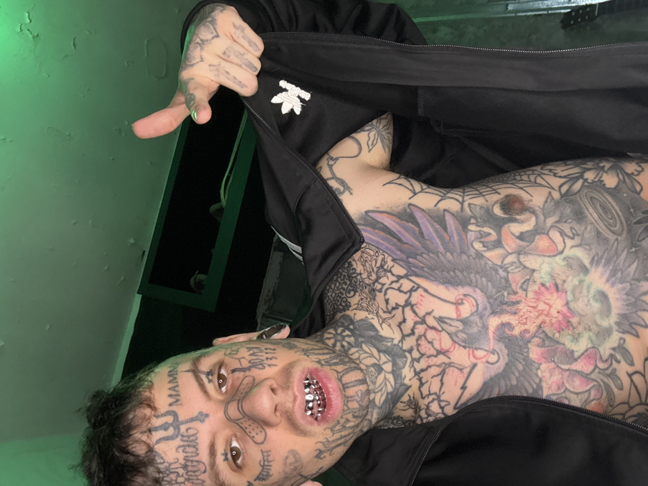 The image depicts a male subject from the chest up, positioned indoors. The subject has extensive tattoos covering his face, neck, chest, and arms. Facial tattoos include a trident symbol on the forehead, the text "MAMN" above the right eyebrow, a band-aid design across the nose bridge, and "AOAX 1312" on the right cheek, among other script and symbols. His hair is dark and styled upwards. The subject has stretched earlobes, with silver-colored tunnels visible. His mouth is open, revealing silver grillz on both upper and lower teeth.
He wears a black tracksuit jacket, open to expose a heavily tattooed chest. A white Adidas trefoil logo is visible on the left chest area of the jacket, and white stripes are partially seen on the shoulder/sleeve. The chest tattoos feature a large central design of a dark bird, possibly a phoenix, with outstretched wings amidst floral and ornamental patterns in various colors, including pink and green. The upper chest area displays a cracked or veined design.
The subject's left hand is raised, with the index finger extended and pointing directly towards the viewer. This finger has visible light green nail polish. The hand and fingers are also extensively tattooed.
The background consists of an indoor wall with visible texture and areas of peeling paint or plaster, particularly on the upper section and ceiling, which are illuminated with a green hue, suggesting a green light source. A dark, rectangular shelf or opening is visible in the upper right background, containing indistinct objects. The specific location within the city of Buenos Aires, Argentina, cannot be determined from the visual information provided in the image.