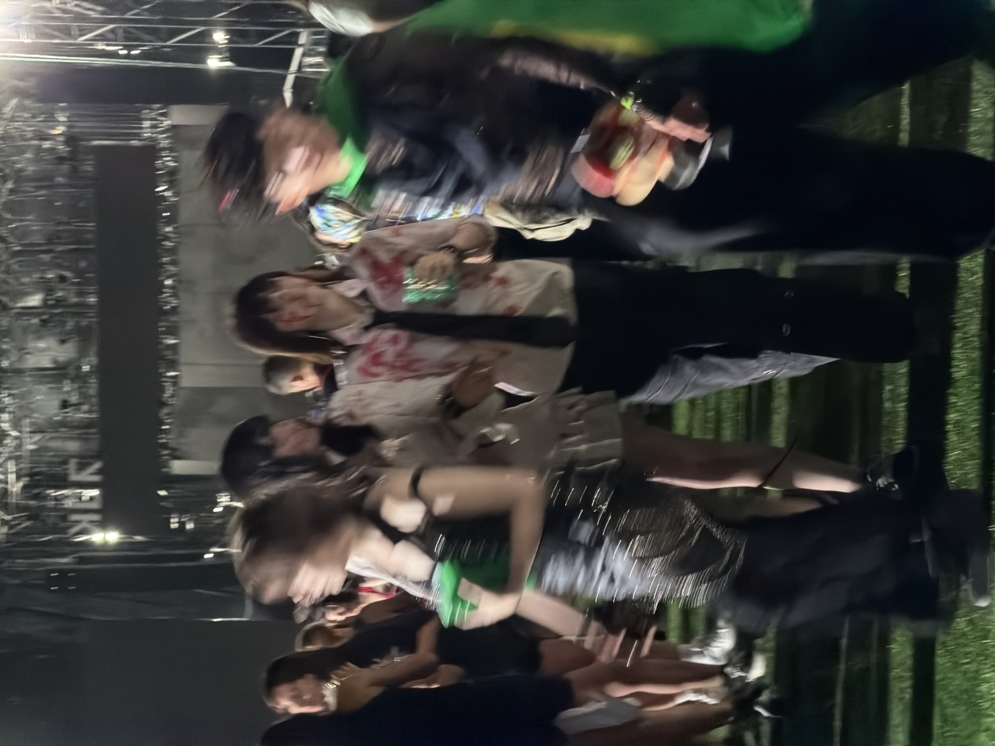 The image depicts a group of young adults outdoors at night, possibly at a concert or festival, given the blurry background which suggests stage lighting and structures. The individuals are dressed in casual, somewhat edgy attire. One person in the foreground is wearing a dark, shimmering top and dark pants, holding a bright green object. To their left, another person has on a white shirt with red splotches, a tie, and dark pants. To the right, a person is wearing a dark jacket over a colorful patterned shirt and a bright green cape-like garment. They are holding a platter with colorful food items. The ground appears to be covered in grass. The overall impression is a candid shot of people enjoying an event in a lively atmosphere, with the lighting creating a dramatic effect. The location is not specified in the image.