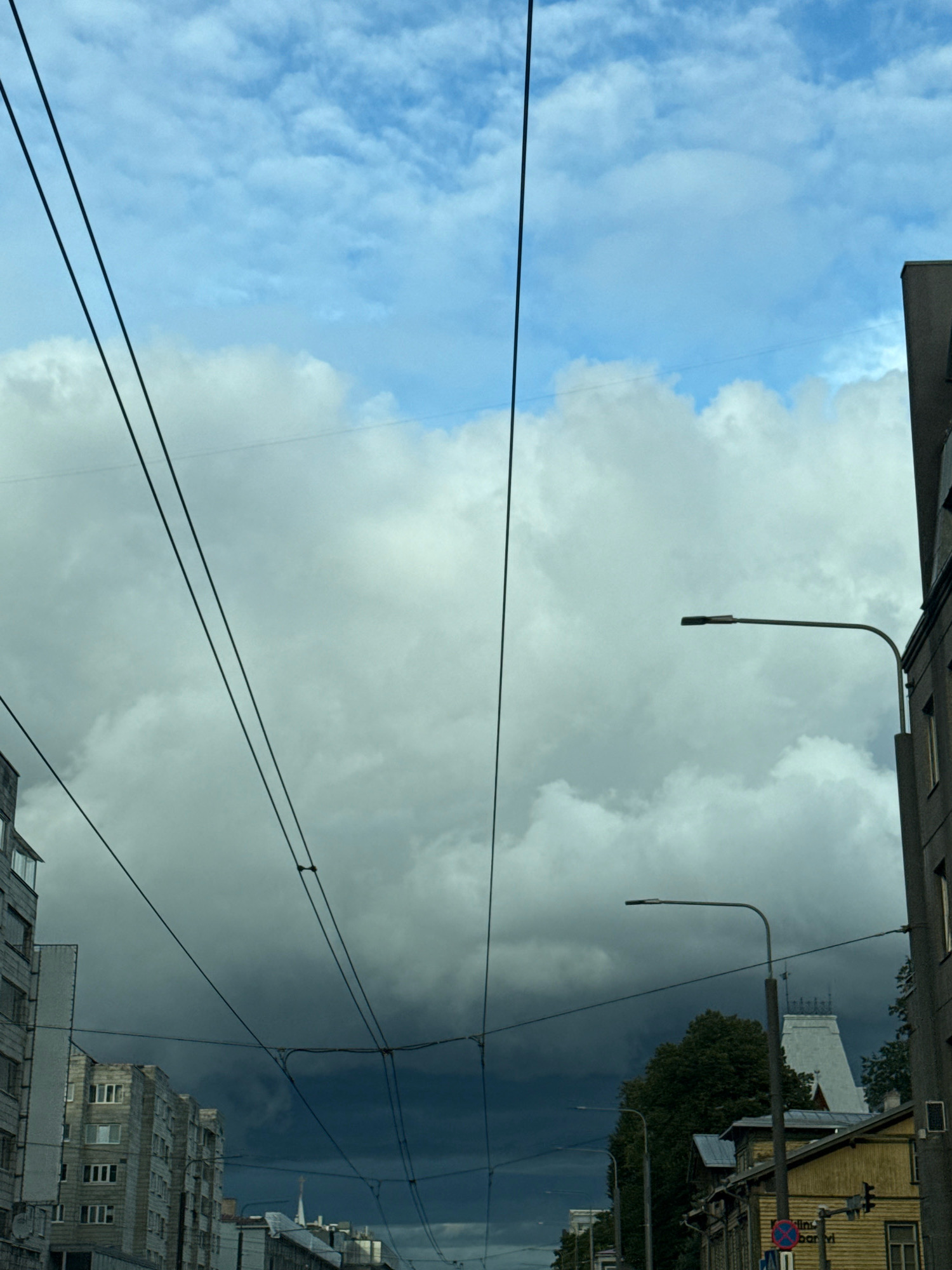 The image captures a street view in Tallinn, Estonia, dominated by a dramatic sky. Thick, dark storm clouds gather overhead, contrasting with patches of lighter, more diffused clouds and a sliver of blue sky in the upper left. Multiple sets of dark overhead wires, likely for trolleybuses or trams, crisscross the frame, adding a linear element against the cloudscape.
In the foreground and mid-ground, elements of the urban environment are visible. On the left, a multi-story apartment building with rows of windows is partially visible. To the right, a modern building facade with windows is present. Further down the street, a cluster of older, lower-rise wooden buildings with yellow siding is seen, one of which has a traffic light attached to a pole. A distinctive white spire or tower-like structure with decorative elements rises above the rooftops. Trees are visible behind the wooden buildings. Two streetlights are also present, one a modern design with a curved arm, and another partially obscured. No people are visible in the frame. The overall impression is one of an approaching storm in an urban setting.