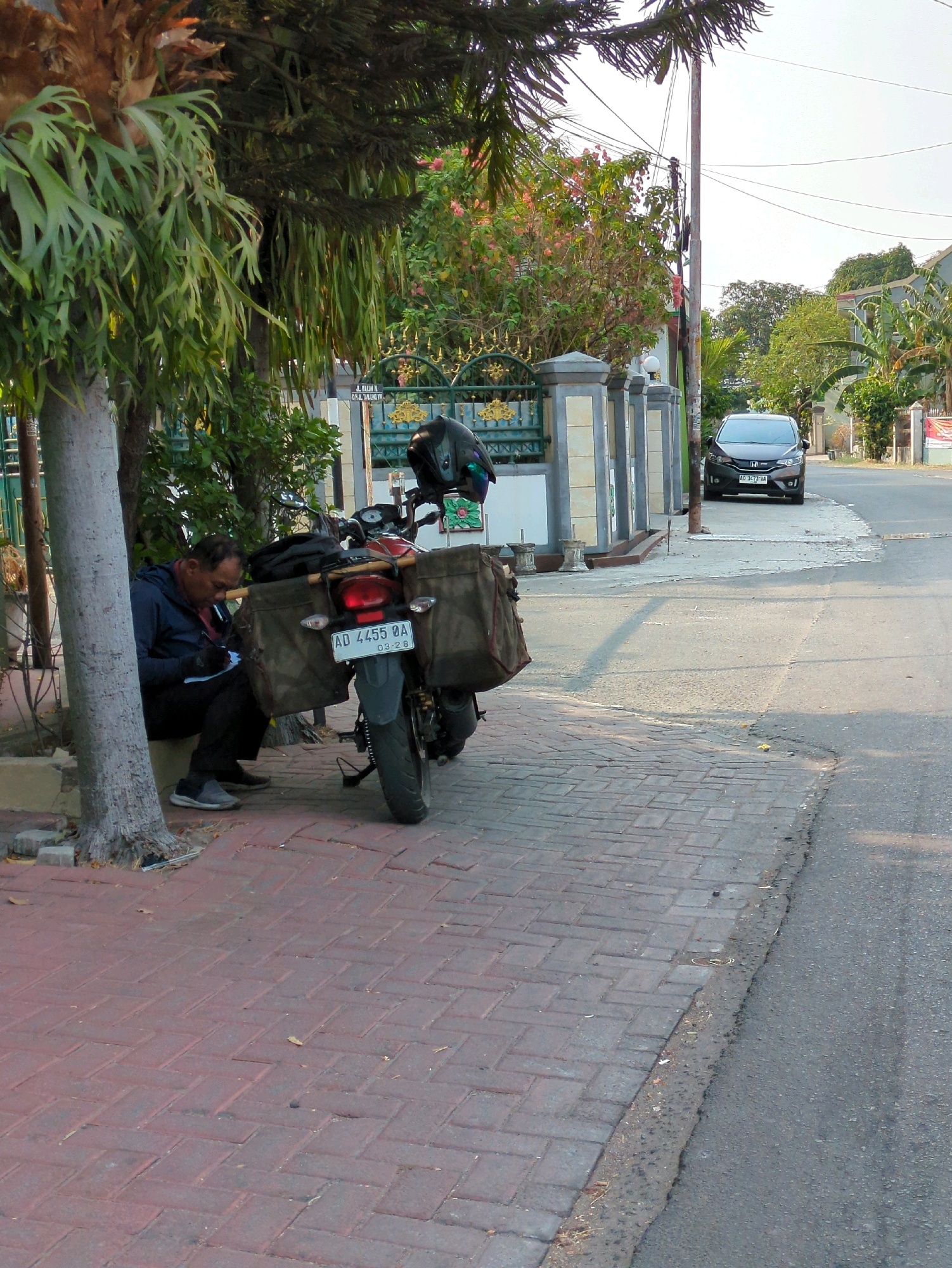 A man sits on a low concrete ledge next to a tree, writing on a piece of paper. He is wearing a blue jacket and black pants. Next to him is a motorcycle with large saddlebags, a helmet perched on the back. The motorcycle has a license plate that reads "AD 4455 QA". The motorcycle and man are on a brick sidewalk next to a street. A dark-colored car is parked on the street in the background. Behind the sidewalk is a fence and a building, and trees line the street. This scene is likely in the city of Surakarta, Indonesia, as suggested by the license plate format and the architectural style of the fence.