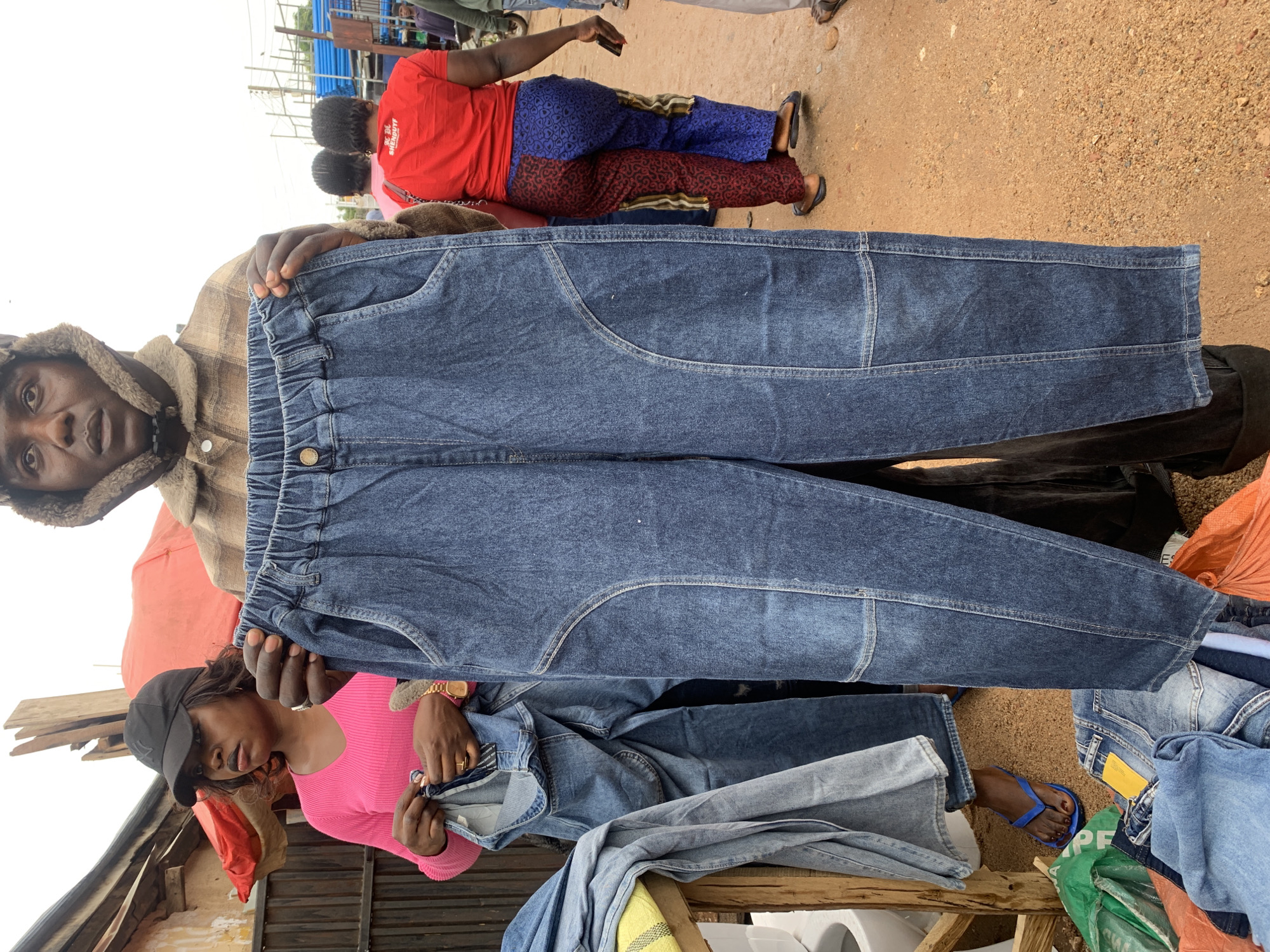 The photo was taken in an open market scene in Gura Top, Nigeria. A man in a shearling-lined hat and tan plaid shirt is holding up a pair of blue denim pants that have an elasticized waist. A woman in a pink shirt and black baseball cap is examining a pair of denim pants. There are several piles of denim pants on a table. In the background, people are standing on the dirt road. There are blue metal pipes stacked vertically, and other goods for sale.