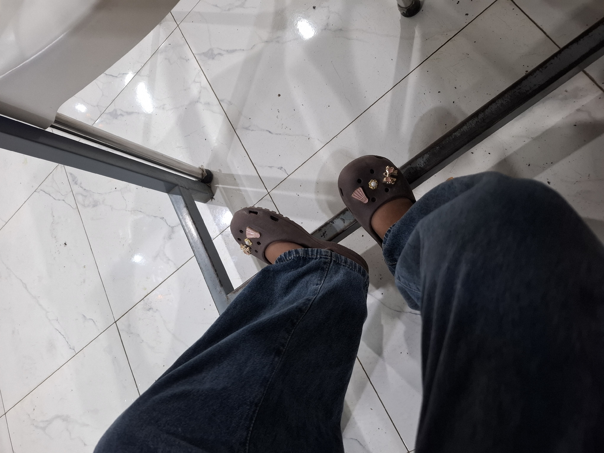 A first-person perspective shot shows a person's legs from the knees down. They are wearing dark blue denim jeans and brown Crocs with decorative charms. The Crocs have charms shaped like a shell, a pearl, and a bow. The person is sitting, as indicated by the angle of their legs and the presence of a metal chair frame. The floor is tiled with white marble-patterned tiles, and there are some dark specks on the floor, possibly dirt. The lighting suggests an indoor environment, potentially a public space or a waiting area. The scene is likely located in Kampongbaro, Indonesia, given the context.